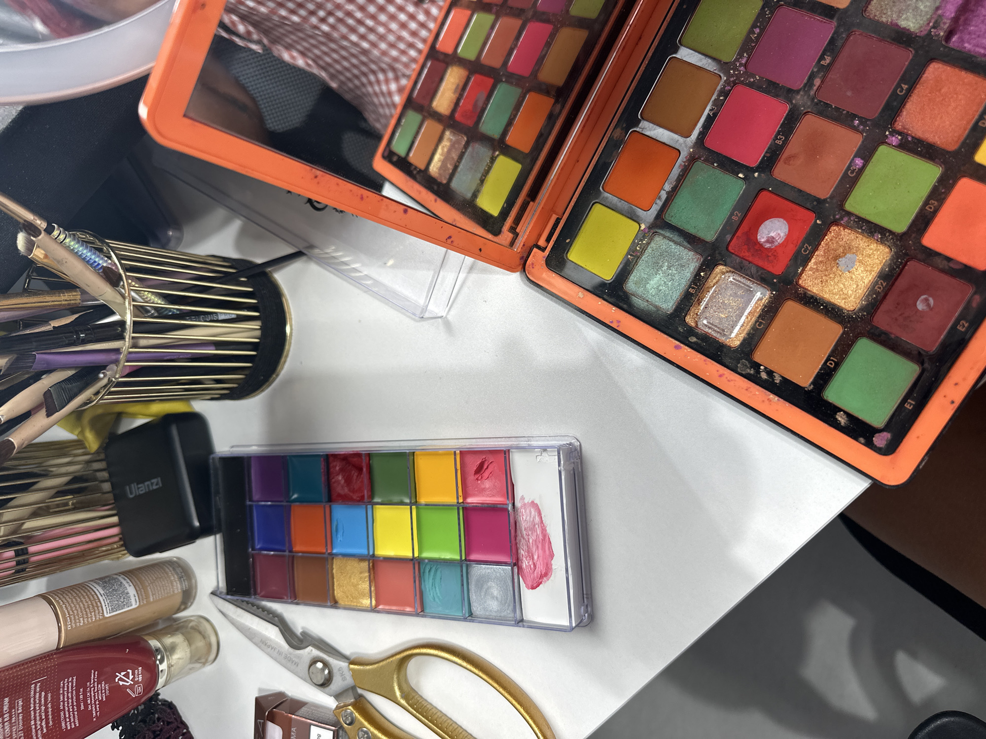 This image shows a collection of makeup items arranged on a white surface, likely a vanity or desk. The subjects are primarily makeup products, including a large eyeshadow palette with many colorful shades, a smaller palette with 24 cream makeup pans, a bottle of BB cream, and a collection of makeup brushes in a gold holder. A pair of gold-handled scissors also lies on the surface, along with a small black electronic device. The eyeshadow palette is open, revealing its contents and some signs of use, with some pans appearing more worn than others. The cream makeup palette also shows evidence of use, with a swatch of pink makeup on a white mixing area. The overall scene is one of a makeup artist's or enthusiast's workspace, with the products neatly organized but clearly in use. The lighting suggests an indoor setting, possibly during the day. Visible text includes brand names like "MISSHA," "Ulanzı," and "SK5," as well as product descriptions and warnings on the BB cream bottle. The location context of North Kuta, Indonesia is noted, though there are no direct visual cues in the image to confirm this specific location.