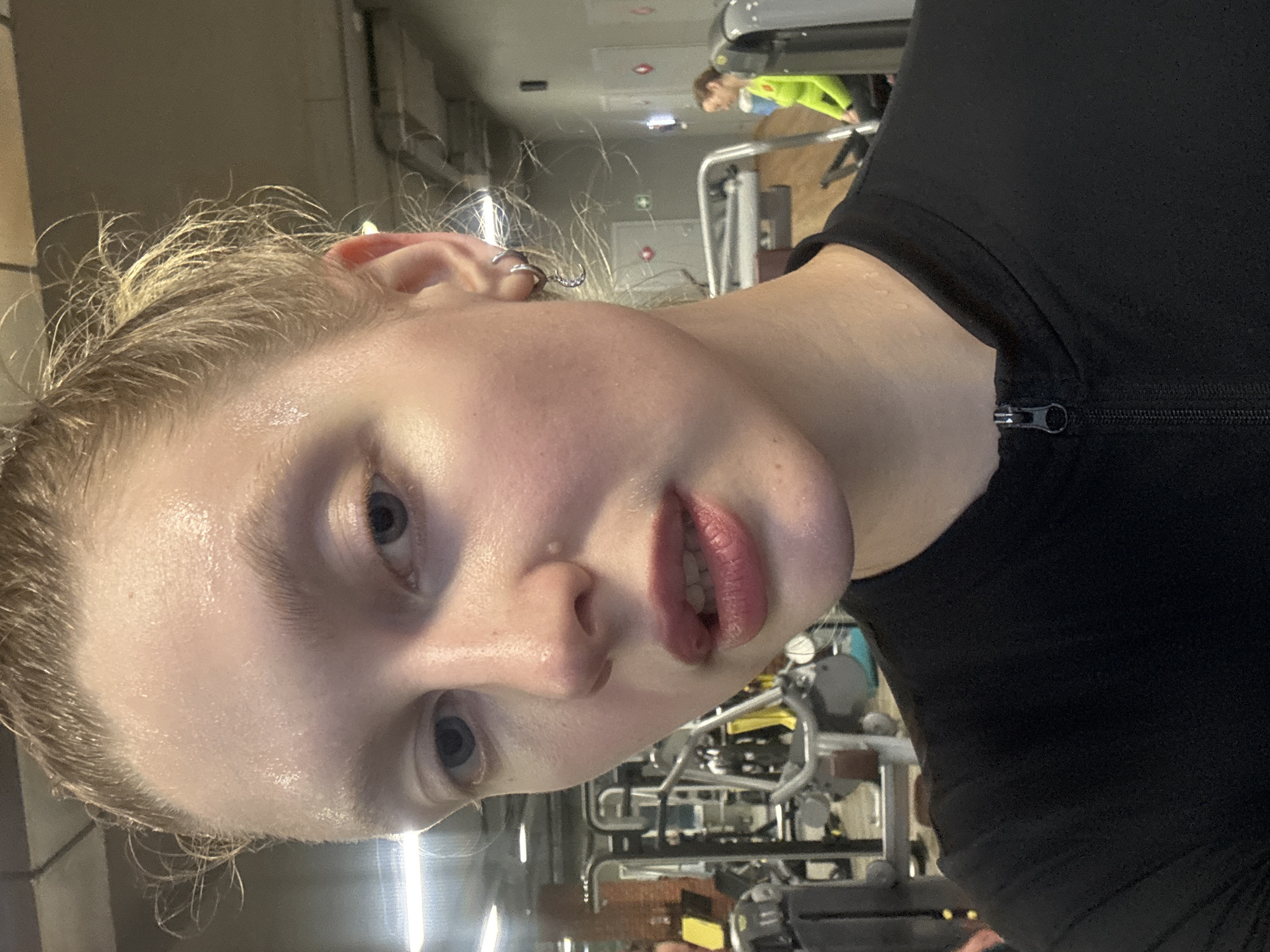 A frontal selfie captures a young Caucasian individual, exhibiting visible perspiration on their face and forehead, situated within a gym in Izhevsk, Russia. The subject has light blue eyes, light-colored eyebrows, and light blonde hair that appears damp and pulled back, with loose strands around the temples. Their lips are slightly parted, revealing a portion of their upper teeth, and possess a natural pink color. They are dressed in a black athletic long-sleeve top featuring a partial front zipper at the collar. The right ear is adorned with two silver hoop earrings, one of which has a small crescent moon charm.
The background clearly depicts a commercial fitness establishment with various exercise machines visible, including multiple resistance training units with black frames and padded seats. The walls are a neutral grey, and the ceiling features exposed ductwork and fluorescent lighting panels. In the far right background, a second individual, a woman in a bright neon yellow-green long-sleeved athletic shirt, is seated on a piece of gym equipment, facing away from the primary subject, engaged in an independent activity. The overall scene suggests a post-workout moment in a modern gym environment.