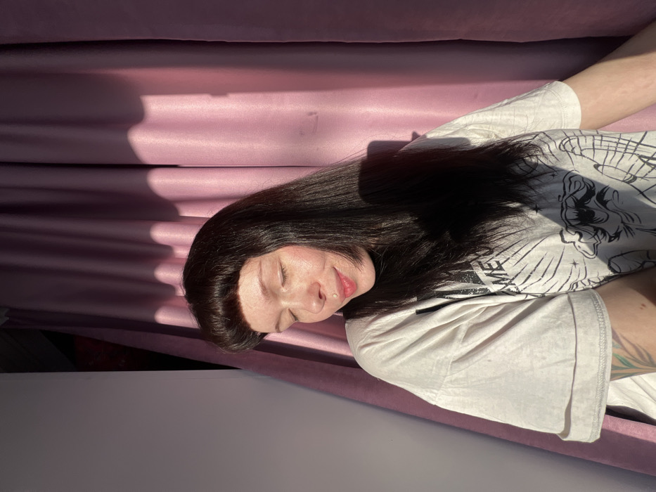 A female subject is depicted indoors, positioned against a light-colored wall on the left and a pleated light purple curtain on the right. She has long, dark hair with blunt bangs, fair skin with visible freckles, and a silver labret piercing on the left side of her upper lip. Her eyes are closed, and her mouth is slightly upturned in a subtle smile. She is wearing an oversized, light-colored t-shirt with a partially visible dark graphic print featuring an illustrative design. A green-toned tattoo is visible on her left forearm. Strong natural light originates from the left, illuminating the left side of her face and upper body while casting portions of her hair and shirt into shadow. Distinct shadows are present on the background curtain. The image provides no visual information to determine the specific location within Chaikovsky, Russia.