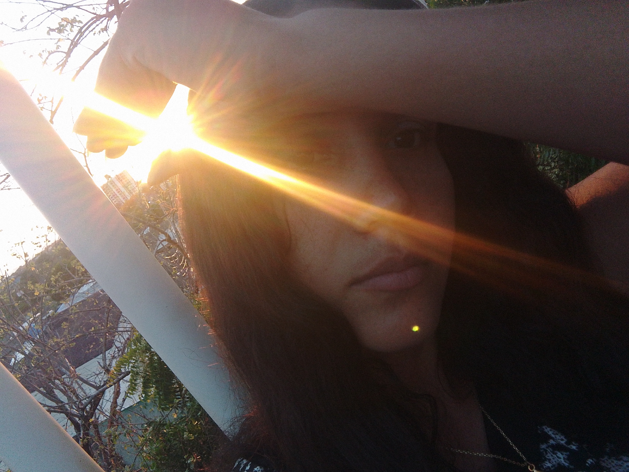 The image depicts a partial, eye-level view of an individual, likely female, in an outdoor setting within Juan Griego, Venezuela. The subject's left forearm is positioned horizontally across the upper face, functioning to block a powerful light source originating from the upper-left corner of the frame. This light source generates a prominent, golden lens flare that extends diagonally across the image. Visible facial features include the right eye, nose, mouth, and chin. Long, dark hair is present. A small, bright yellow-green reflective point is discernible on the right cheek or jaw area. A gold-colored chain necklace with a pendant is visible near the lower right. The background, partially framed by two white diagonal bars, shows an outdoor environment containing trees with both bare branches and green foliage, along with indistinct structures, including a dark-colored object resembling a vehicle and a building. The scene is under strong daylight conditions.