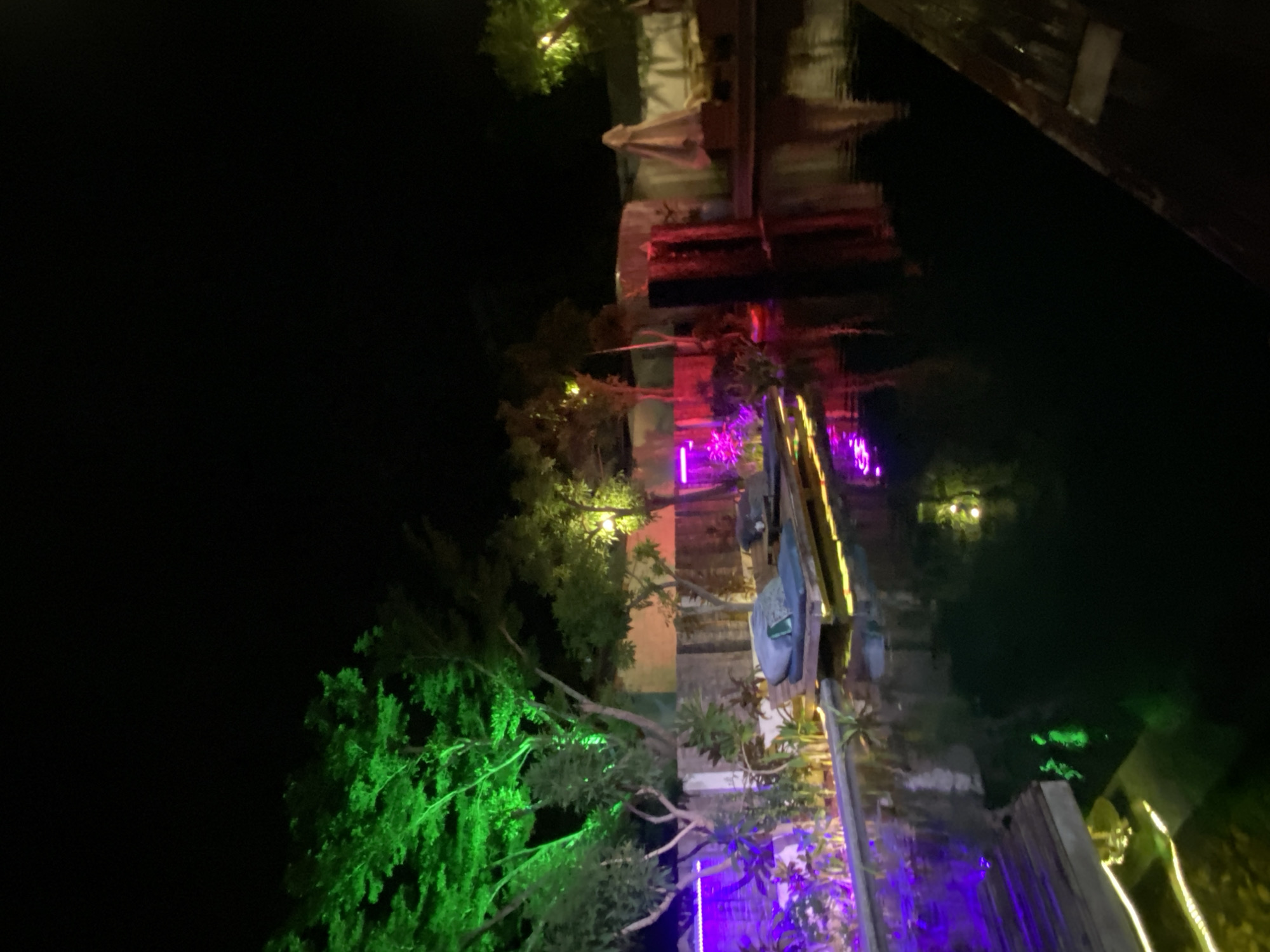The image depicts a nighttime scene in Mũi Né, Vietnam. A body of water reflects colorful lights illuminating surrounding vegetation and structures. Green light shines on trees in the foreground, while purple and red lights highlight buildings and other elements. A wooden structure is visible on the right side, and a small boat is reflected in the water. The background is largely dark, emphasizing the illuminated elements.