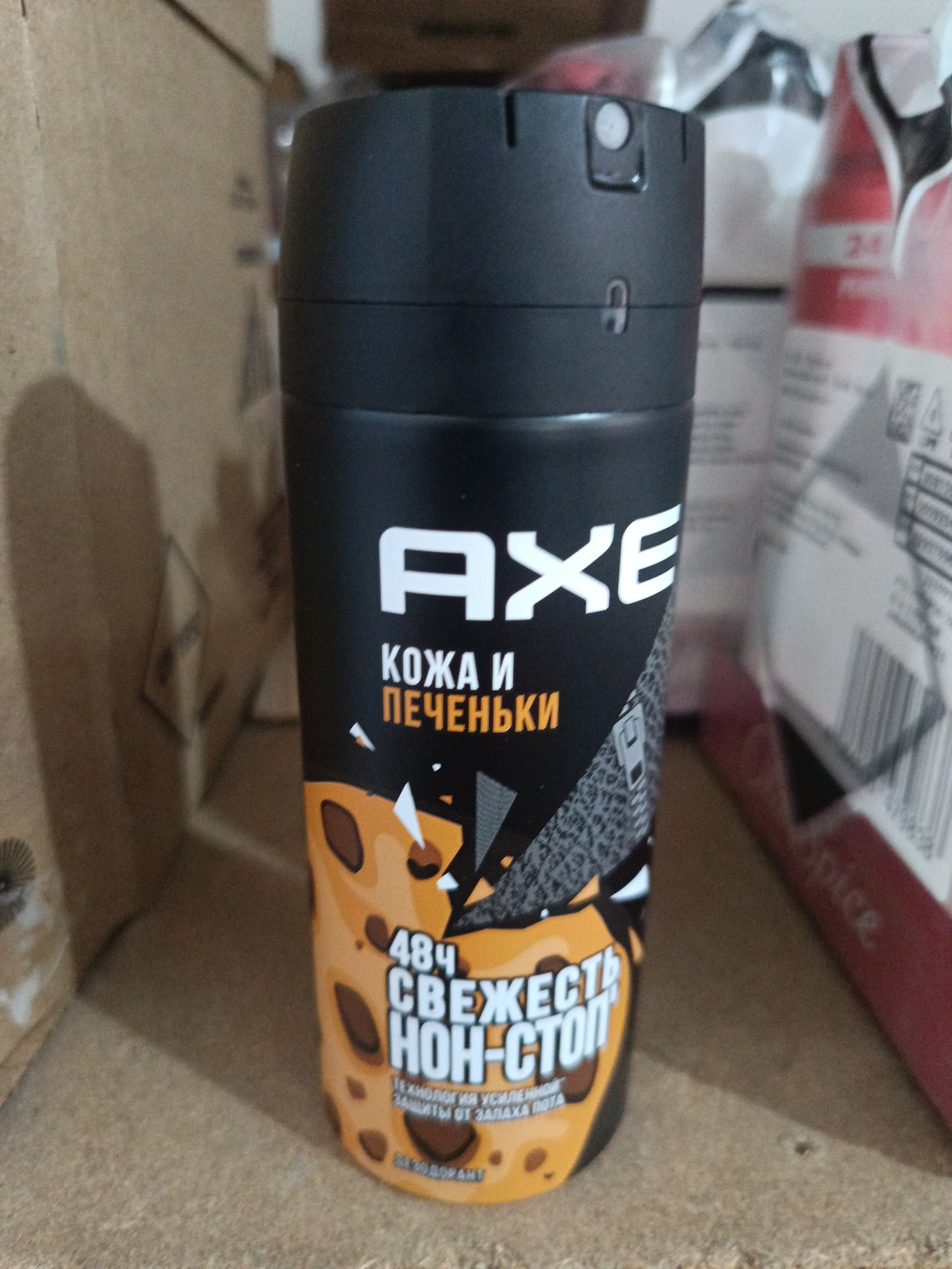 The image shows a black spray can of AXE deodorant with a yellow and brown leopard print design. The can is placed on a wooden surface, with cardboard boxes in the background. The text on the can is in Russian and includes "AXE," "КОЖА И ПЕЧЕНЬКИ" (skin and cookies), and "484 СВЕЖЕСТЬ НОН-СТОП" (484 freshness non-stop). The context of Berdiansk, Ukraine, is not visually represented in the image.