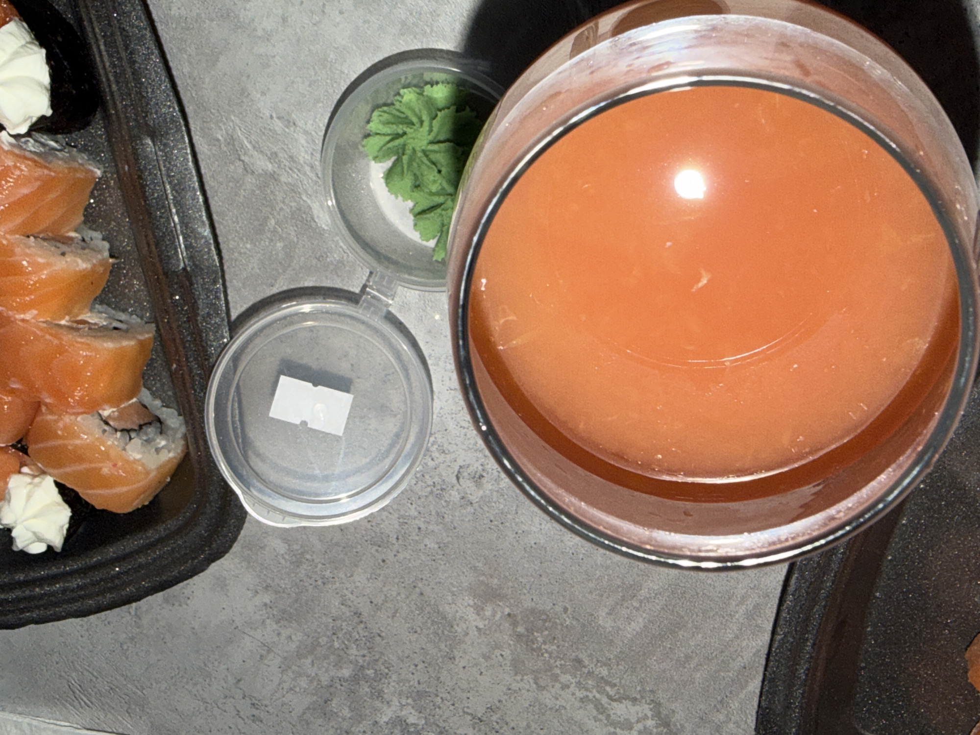 An overhead shot captures a casual meal featuring sushi and a drink on a gray, textured surface, likely a table or countertop. The primary subjects are a black rectangular tray filled with sushi rolls, a large glass containing an orange beverage, and two small clear plastic containers.
On the left, several sushi rolls are arranged in the black tray. These appear to be salmon rolls, possibly Philadelphia rolls, with bright orange salmon layered over white rice and a dark seaweed wrapper, and some rolls topped with dollops of white cream cheese. Next to the sushi, a small clear plastic lid is open, revealing a small, white, rectangular object inside, which could be a packet or a piece of food.
To the right of this, another small clear plastic container holds a generous portion of green wasabi, shaped decoratively. Dominating the right side of the frame is a large, clear glass filled with a vibrant orange liquid, possibly juice or a cocktail. A bright reflection is visible on the surface of the drink, suggesting an overhead light source.
The scene suggests an indoor setting, possibly a home, as indicated by the takeout-style containers. The lighting is somewhat dim, creating reflections on the glass and casting soft shadows. No people are visible, and there is no discernible text on any of the items. The overall impression is one of a prepared meal ready for consumption.