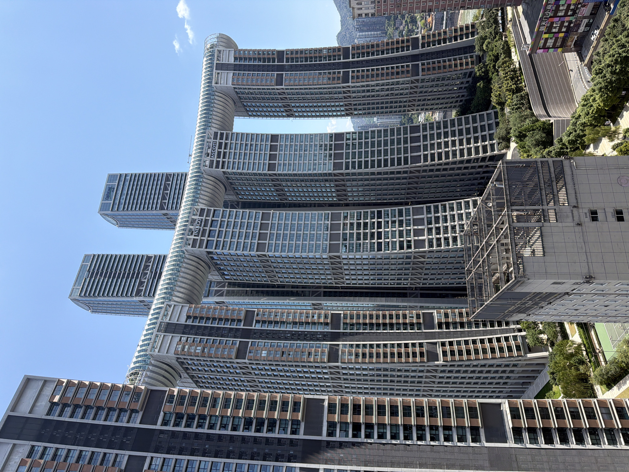 The image showcases the unique architecture of the Raffles City Chongqing complex in Yuzhong District, China. The central focus is the iconic "Crystal" structure, a horizontal skybridge connecting four of the eight skyscrapers. The skybridge features a transparent, curved roof and is supported by arch-like structures that bridge the gaps between the towers. The skyscrapers themselves are modern, high-rise buildings with numerous windows and varying facade designs, primarily in shades of gray and beige with some reddish-brown accents.
The surrounding environment includes other tall buildings, some with distinctive architectural features like the one on the far left with its dark, textured facade and repeated balcony structures. In the foreground, a portion of another building is visible, featuring a concrete rooftop with metal railings and a grid-like structure. Below the skybridge and between the towers, lush green trees and landscaped areas are visible, creating a contrast with the dense urban development. A tennis court is also discernible in the lower left section of the scene, surrounded by greenery. Further in the background, a river can be seen, with hilly terrain and more buildings on the opposite bank. The sky is clear and blue with a few wispy clouds. The overall impression is a densely built urban landscape characterized by impressive and innovative architecture.
