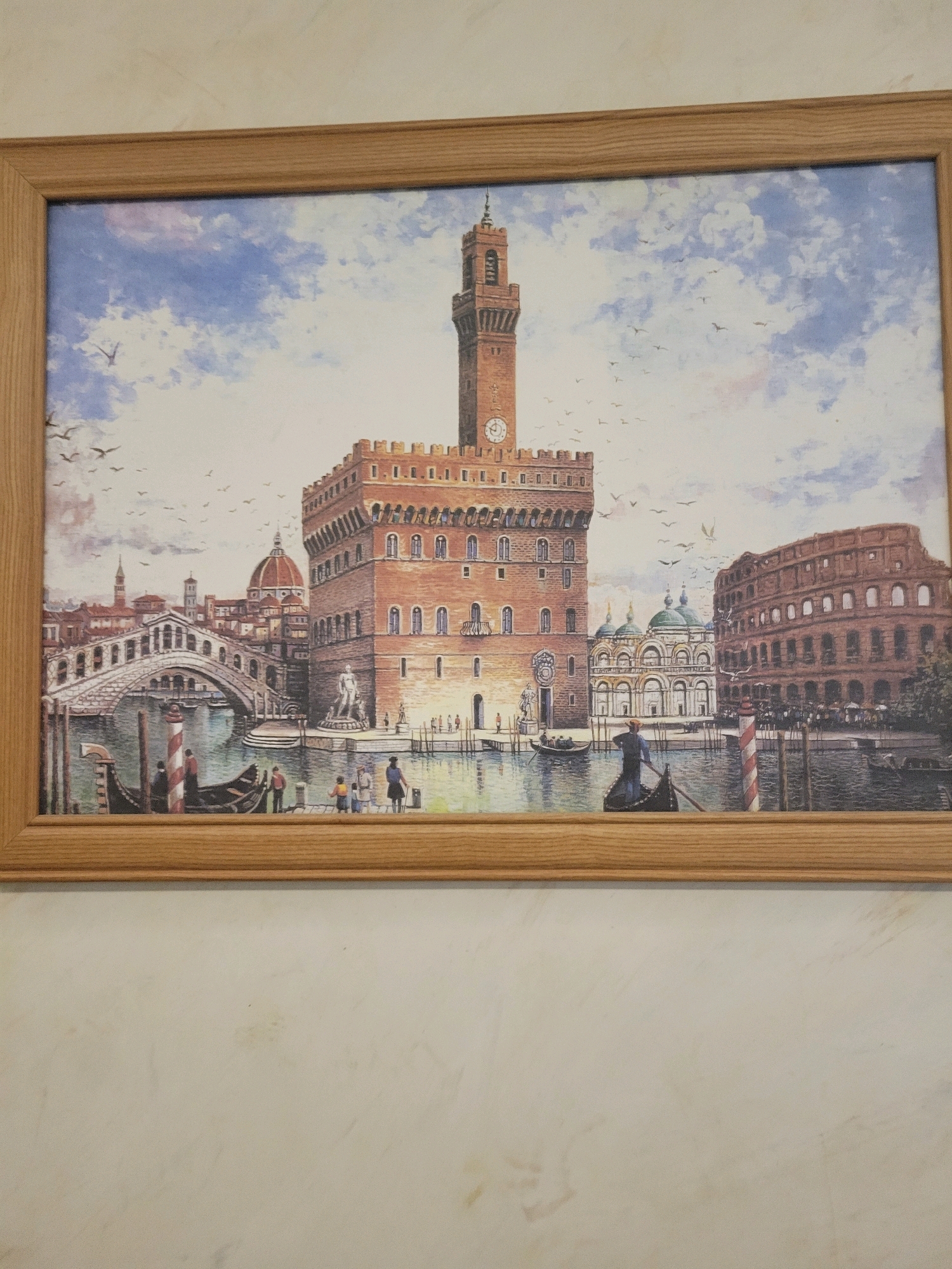 The image shows a framed painting of a European cityscape. The painting depicts a canal with gondolas, a large brick building with a clock tower, a domed building, and an arched bridge. There are several figures in the painting, some in gondolas and some standing on the shore. In the background, there are more buildings and a cloudy sky with birds. The painting is hanging on a light-colored, textured wall. The wall has some subtle stains or marks on it. The context provided mentions Sachkhere, Georgia, but the painting itself depicts a scene that is not identifiable as Sachkhere, Georgia.