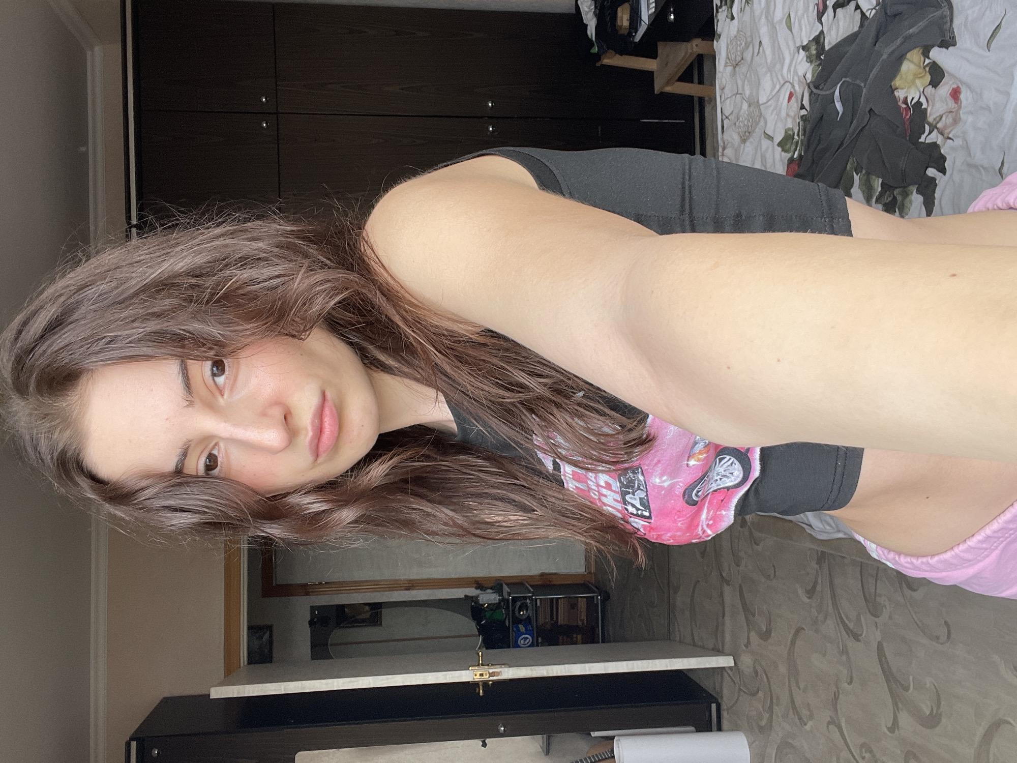 A young woman with long, wavy brown hair and fair skin is taking a selfie in an indoor setting. She is wearing a black top with a pink and white graphic design, and pink shorts. The background features a dark brown wardrobe with silver handles, and a bed with a floral patterned bedspread. There is also a cabinet with items on it visible in the background. The location cannot be identified as Moscow, Russia from the image.