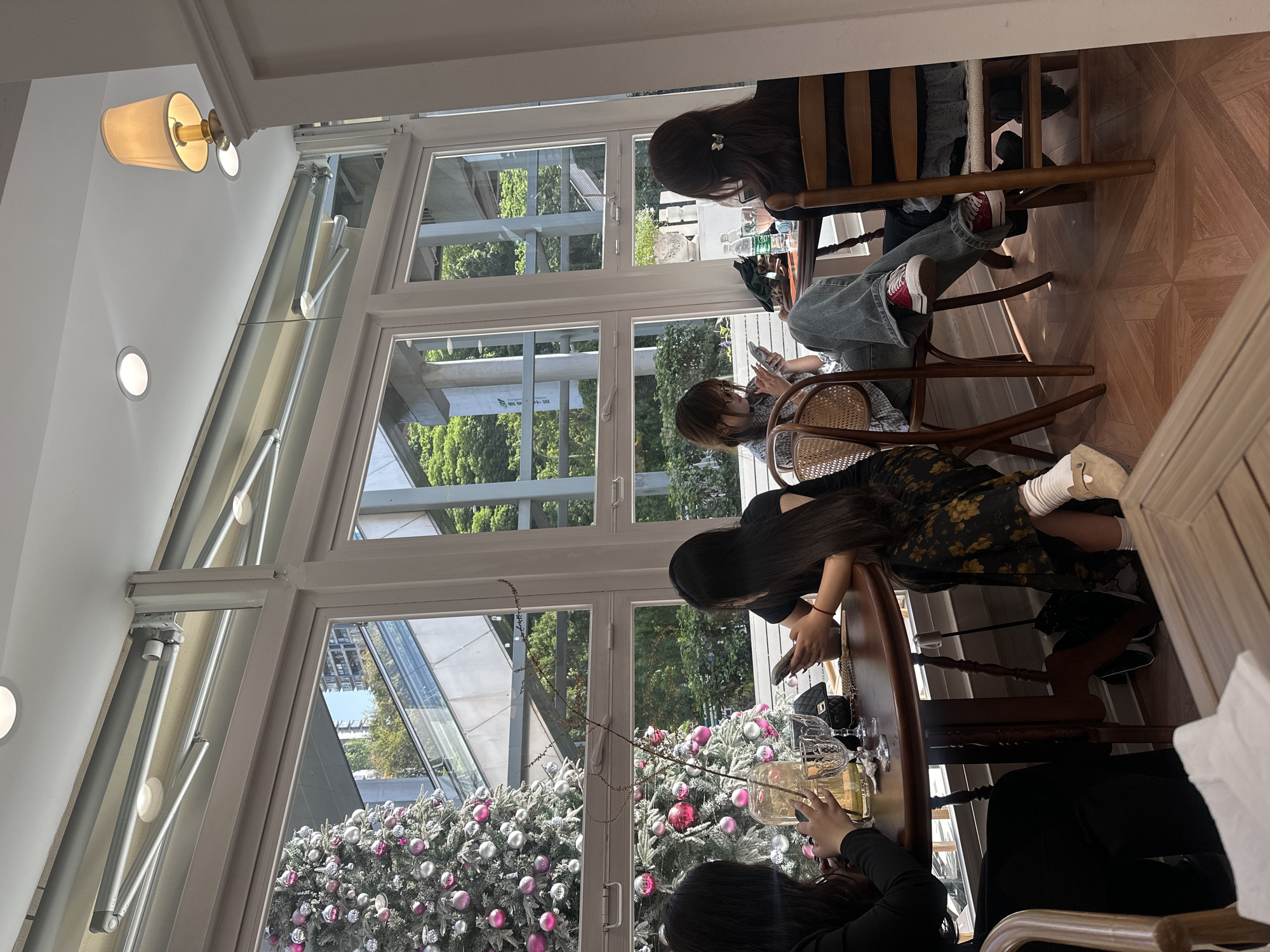 The image depicts a group of four people sitting at a table in what appears to be a café or restaurant, judging by the tables, chairs, and glassware. The location is characterized by large windows providing an exterior view. Outside the windows, decorated Christmas trees are visible. The individuals are engaged in different activities: one person is focused on a mobile phone, another is looking down at the table, and the others are oriented towards the objects on the table. The flooring is wood, and the lighting is soft, provided by ceiling lamps. Given the context of China, and the likely presence of Christmas decorations, this could likely be a western-style cafe. Given that Liangjiang is a special economic zone in Chongqing China, the building is very likely inside a commercial structure within the administrative area.