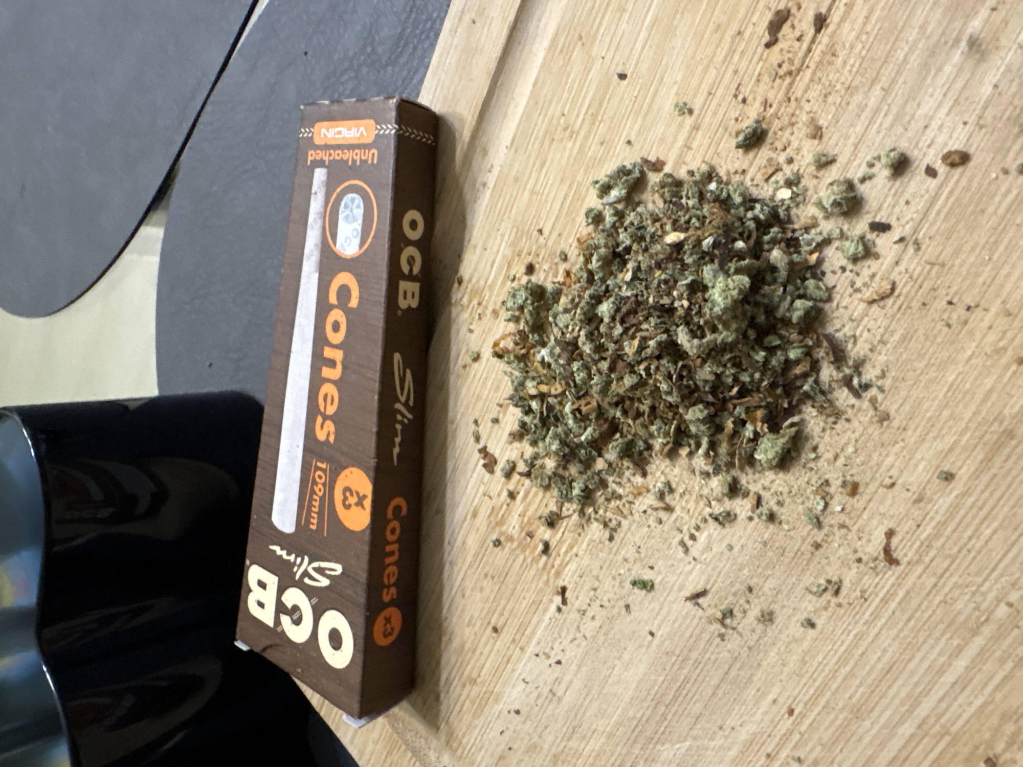 The image shows a wooden surface upon which is a pile of what appears to be marijuana and a box of OCB Slim Cones (Unbleached Virgin, 109mm). A black object is partially visible to the left. The location is not identifiable in the image.