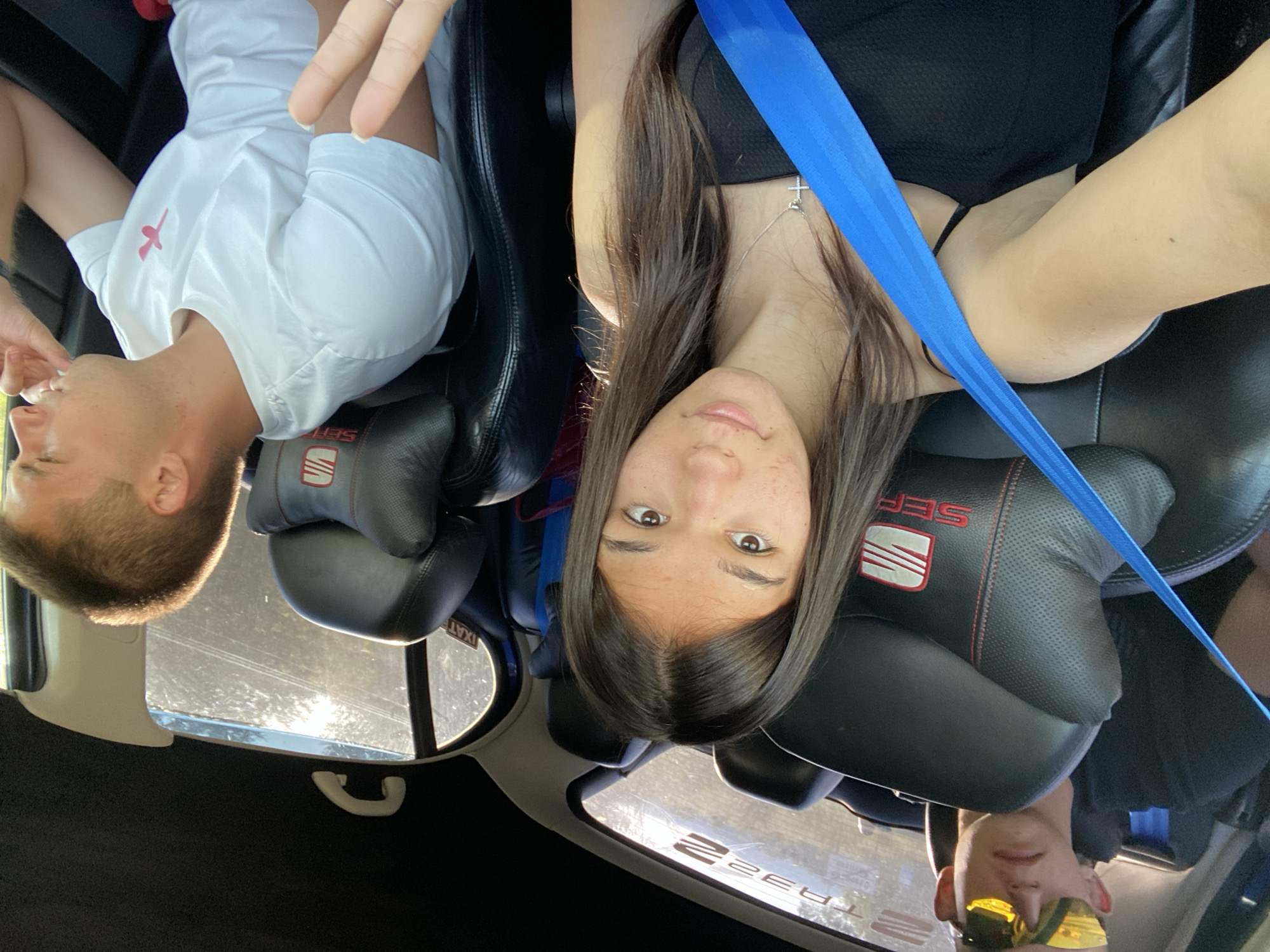 In the city of Sevlievo, Bulgaria, three individuals are seated inside a car. The person in the foreground, a young woman with long brown hair, is taking a selfie. She is wearing a black sleeveless top and a silver necklace with a cross pendant. A bright blue seatbelt crosses diagonally across her body. To her left and behind her, partially visible, is a young man wearing sunglasses and a black t-shirt. To her right and further back, another young man is seated, wearing a white t-shirt with a pink logo. The interior of the car features black leather seats with red stitching, and the brand "SEAT" is visible on the headrests. The scene suggests a casual outing or journey.