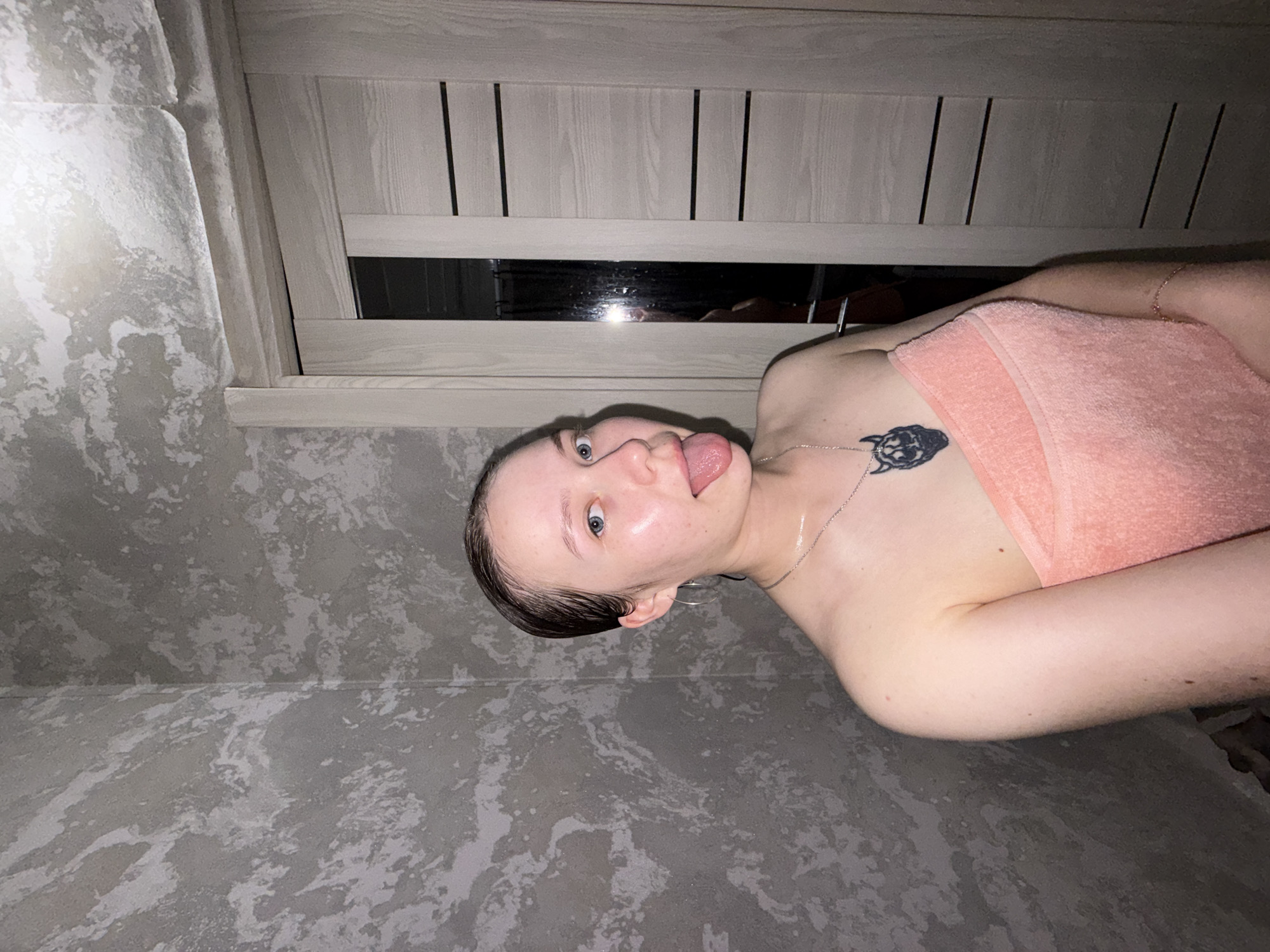 A woman with wet hair is sticking her tongue out, wearing a towel, and posing in what appears to be a bathroom with gray, mottled walls and a wooden-slatted window above. She has a tattoo on her chest and is wearing a necklace. The scene is dimly lit. The location is potentially Chaikovsky, Russia.