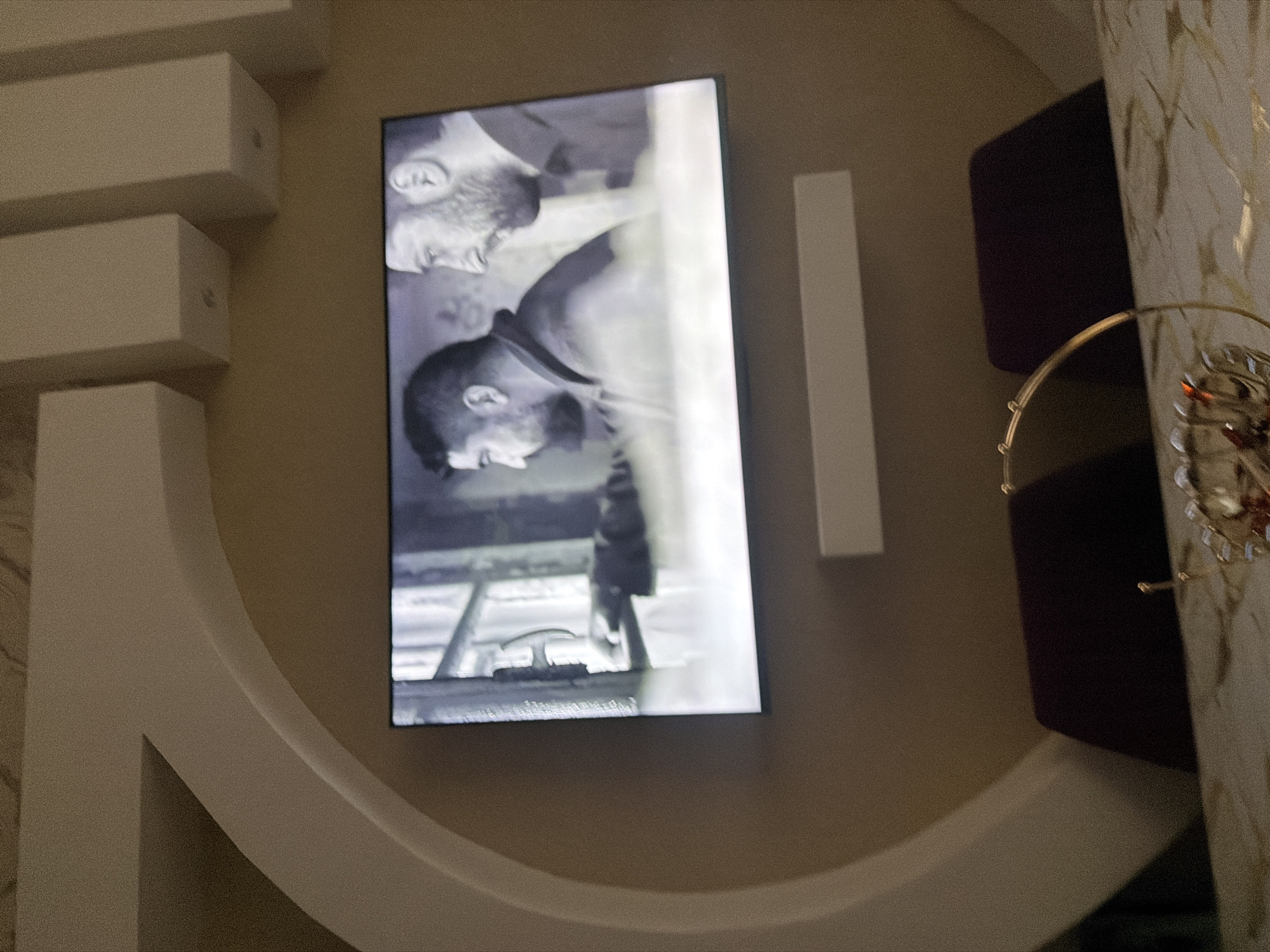The image captures a modern living room setting, likely in the city of Digah, Azerbaijan. A large flat-screen television is centrally mounted on a beige wall, displaying a black and white scene from a film. On the screen, two men are visible, one in the foreground facing right with a beard and short dark hair, and another behind him facing left, also with short dark hair. The man in the foreground appears to be looking at something to his right.
The television is positioned within a large, circular, beige architectural element that protrudes from the wall. Above the television, three rectangular white structures are mounted on the wall, possibly decorative elements or lighting fixtures. Below the television, a long, narrow, horizontal white shelf or fixture is mounted.
In the foreground, a table with a marble-like surface is partially visible. On the table, a golden decorative piece with a circular frame and several small spheres is present. The overall lighting is dim, suggesting it is either evening or the room's lighting is subdued. Two dark-colored chairs are visible behind the table, with their tops extending into the frame. The wall and architectural elements are painted in neutral, light beige tones.