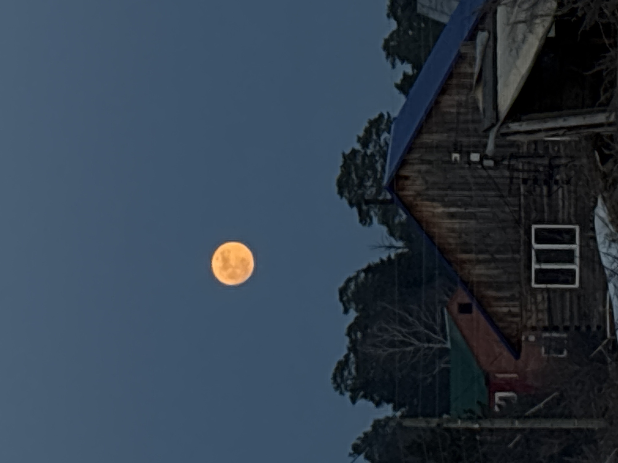 The image shows a full moon in a dark blue sky. To the right of the sky, there is a wooden house with a blue roof. There are trees behind the house. The image appears to be taken at night. Based on the image, it is not possible to determine the location in Chaikovsky, Russia. More details are needed to pinpoint a specific place within the city.