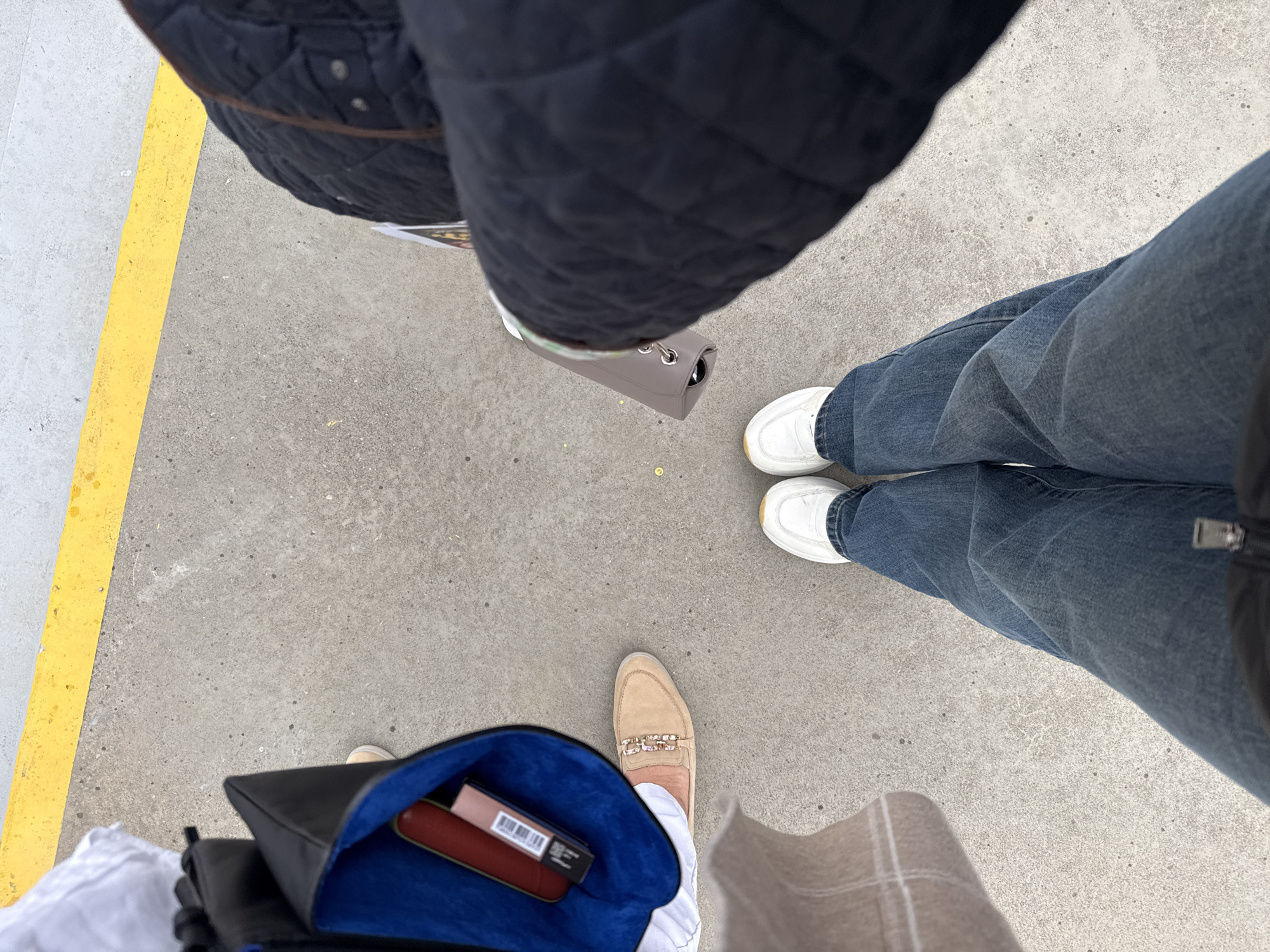 The image depicts a view looking down at three people standing on a concrete surface with a yellow line on the left edge. The person on the top is wearing a navy quilted jacket and carrying a gray handbag. The person on the right is wearing jeans and white sneakers. The person on the bottom is carrying a black bag with a blue interior containing a lipstick and other cosmetic items. The person is also wearing tan-colored loafers with decorative embellishments and beige trousers. The location is suggestive of an urban environment. There is no mention of the city of Sofia, Bulgaria.
