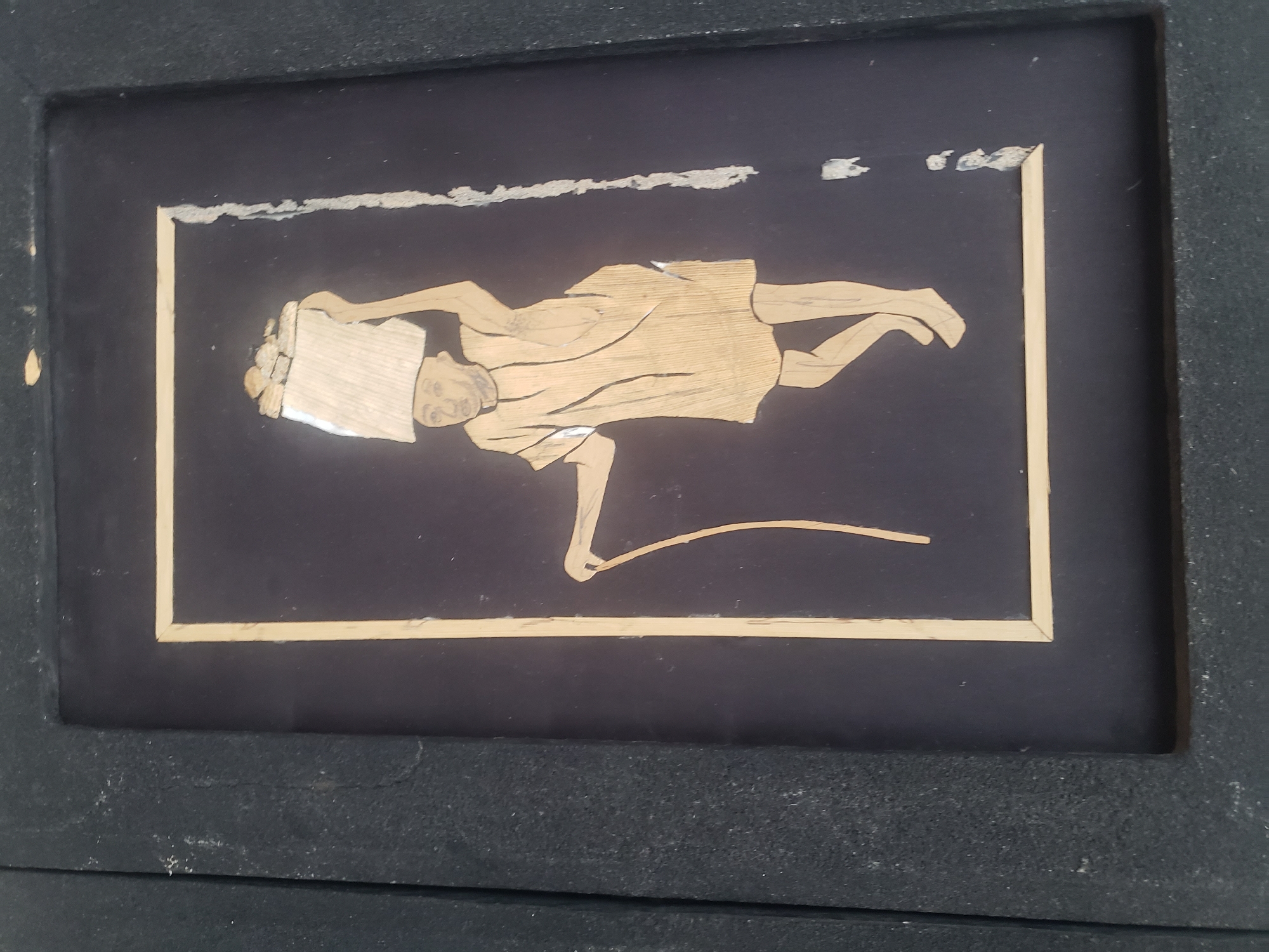 The image is a close-up, straight-on view of a framed artwork. The artwork itself is a silhouette of a person carrying a large sack or basket on their head, with a cane in their left hand. The figure is rendered in a light-colored material, possibly straw or wood, against a dark background. The frame appears to be black and textured. The artwork is positioned within a larger, dark, textured border, suggesting it's part of a larger display or piece of furniture. The image's focus is on the artwork and its immediate surroundings. The specific location in Sabon Tasha, Nigeria is not discernible from the visual information provided in the image.