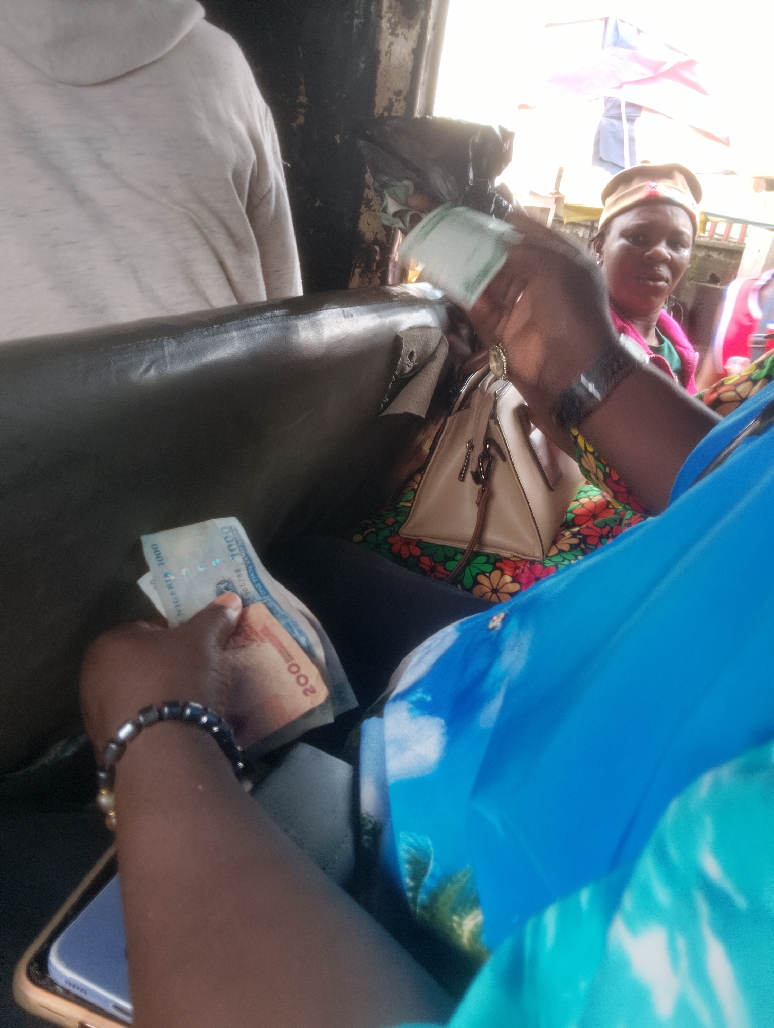 In Ikeja, Nigeria, a close-up shot shows a person's hands holding Nigerian currency, specifically what appears to be a 1000 Naira note and a 200 Naira note. The hands, adorned with a beaded bracelet, are positioned over a dark leather seat, possibly in a vehicle. In the background, another person wearing a cap and a green shirt is visible, partially obscured by the foreground subject's arm and a tan handbag. The setting appears to be a bustling urban environment, suggested by the bright, natural lighting and glimpses of what could be market stalls or other outdoor structures. The overall scene depicts a moment of financial transaction or handling of money within the lively atmosphere of Ikeja.