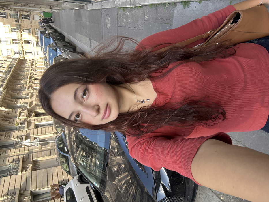The image is a selfie of a young adult female positioned centrally in an urban streetscape in Paris, France. The female subject has long, wavy reddish-brown hair and a fair complexion, wearing a rust-colored, ribbed, long-sleeve top and a silver-toned necklace with multiple small dark charms. She holds the camera with her right arm extended, with a brown leather shoulder bag partially visible on her right side.
Behind her, a dark-colored sedan is parked on the street, its front grille and windshield reflecting the surrounding buildings. The street extends into the background, lined with a continuous row of parked vehicles. On both sides of the street, classic Haussmannian-style buildings with light-colored stone facades, multiple stories, ornate window frames, and wrought-iron balconies are visible. A pedestrian walks in the distance on the right sidewalk, near a green public waste bin. The scene is illuminated by natural daylight, casting clear shadows and highlighting the architectural details.