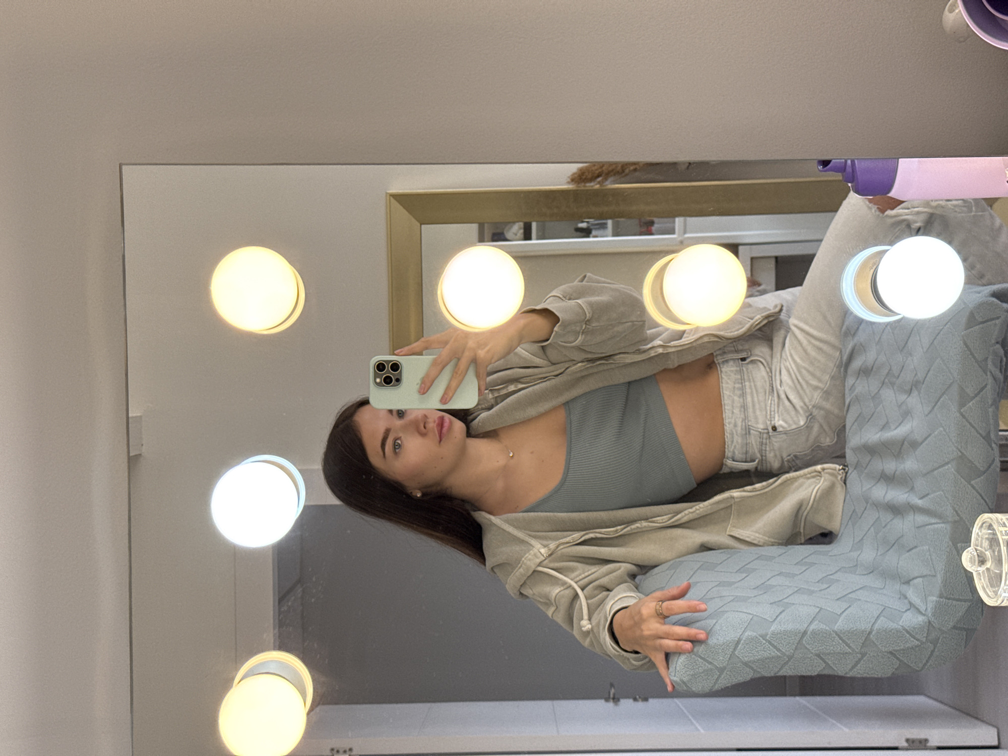 A young woman is taking a mirror selfie in what appears to be a bathroom or vanity area. She is holding a smartphone in her left hand, angled toward the mirror to capture her reflection. She has long brown hair, is wearing a light-colored zip-up hoodie, a grey ribbed top, and faded jeans. She is partially reclined on a large textured cushion. The mirror has vanity lights around it, with several bulbs illuminated. Other items visible in the background reflection include toiletries and possibly additional shelving. The image provides no definitive information to accurately place the location within Ramat Gan, Israel.
