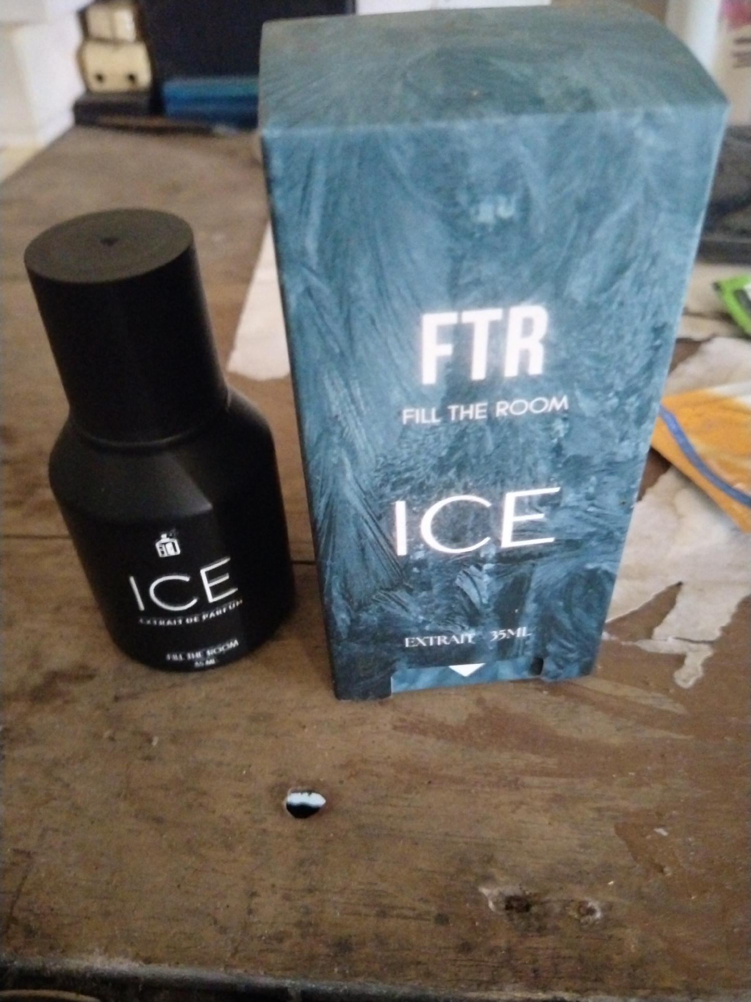 A black bottle of "ICE" perfume and its matching teal-colored box, featuring a frosty pattern and "FTR FILL THE ROOM ICE" text, are placed on a weathered wooden surface. The bottle has a black cap and text indicating "EXTRAIT DE PARFUM" and "FILL THE ROOM 45 ML". The box also displays "EXTRAIT 35ML". The background is blurred, showing hints of light-colored objects. The scene is devoid of human presence. The image appears to be taken indoors, possibly in a domestic setting. There is no visual information available to confirm the location in Tegaldowo, Indonesia.