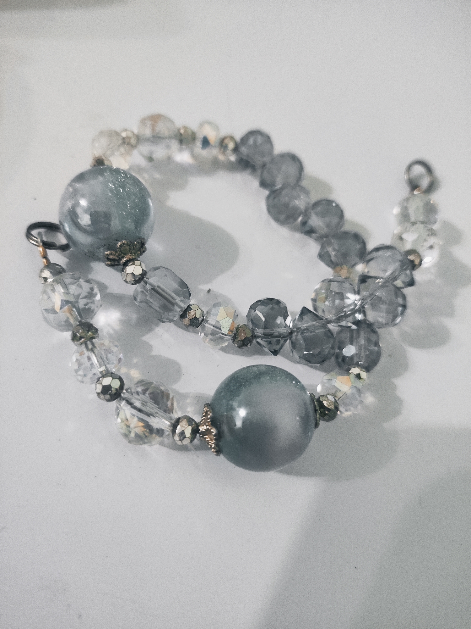 The media file shows a close-up of a beaded bracelet against a white background. The bracelet is made of two strands. One strand consists of alternating clear faceted beads and opaque gray beads. The other strand features mostly opaque gray faceted beads. There are also smaller, metallic beads used as spacers. Two larger, smooth, opaque gray spheres are prominent features on the bracelet. One sphere is near the top left, and another is towards the bottom right. The lighting creates shadows on the white surface, suggesting an indoor setting. There is no text visible in the image. The overall impression is that of a piece of jewelry displayed on a flat surface.