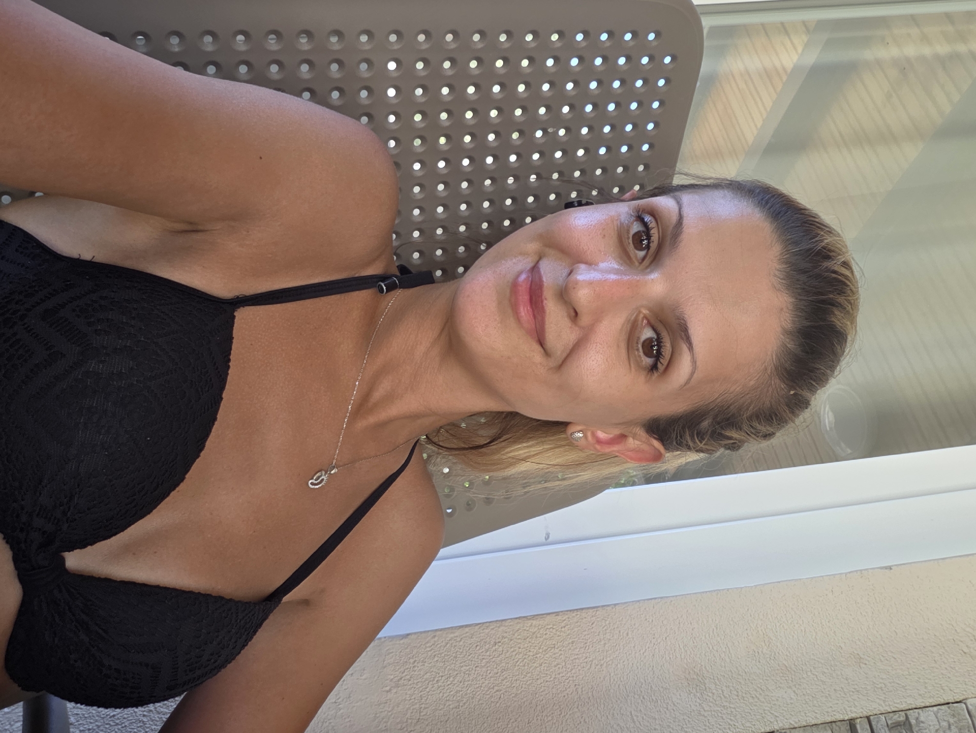 A woman with blonde hair pulled back and wearing a black bikini is seated in a lounge chair, looking at the camera and smiling. She is wearing a silver necklace with a pendant and small stud earrings. She has sunglasses in her hair and what appears to be a black earbud in her left ear. The background shows a white frame structure and textured wall, consistent with an outdoor patio or balcony area. The location is the city of Omiš, Croatia.