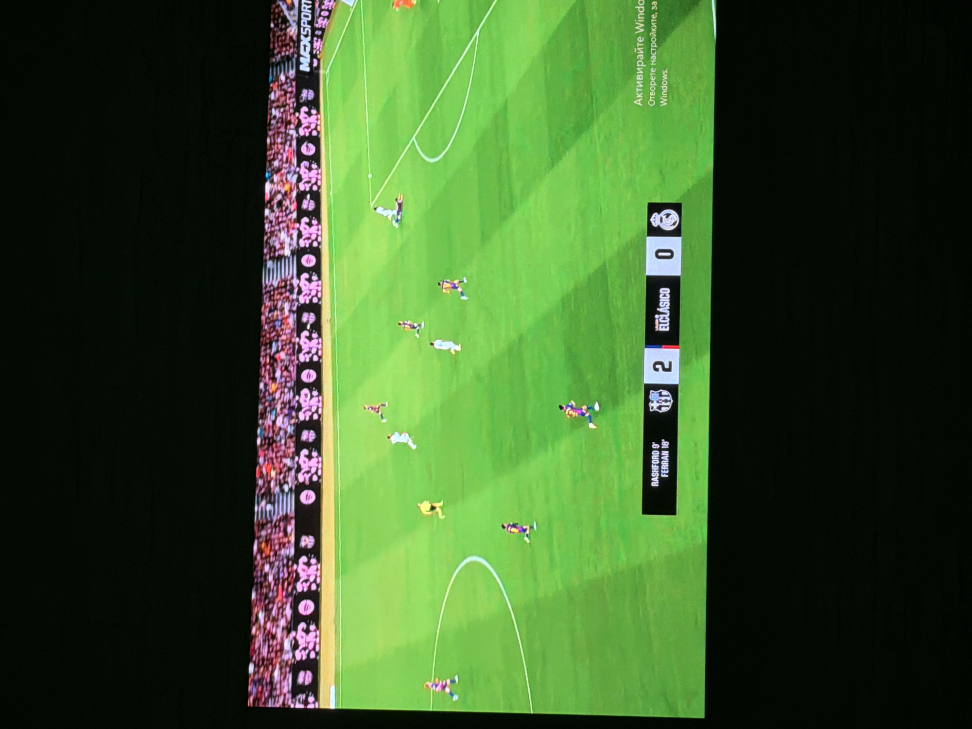 The image shows a soccer game, specifically "El Clásico," where Barcelona is playing against Real Madrid. The score is 2-0 in favor of Barcelona. The goals for Barcelona were scored by Rashford in the 9th minute and Ferran in the 18th minute. The game is being displayed on a screen, potentially a television or monitor. The surrounding environment is dark, indicating it's an indoor setting. The edge of the screen also shows "MAXSPORT" sponsorship branding. The Windows operating system on the screen is not activated, as indicated by the "Activate Windows" message.
