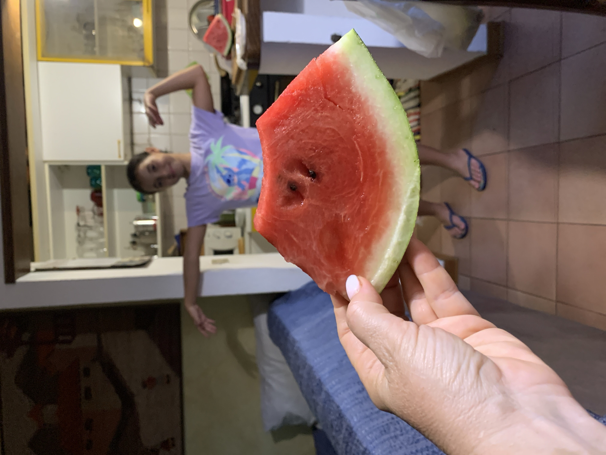 The image shows a close-up of a person's hand holding a slice of watermelon in what appears to be a kitchen. In the background, a young person in a purple t-shirt is posing. The kitchen has white cabinets and tiled floors. The setting suggests it is daytime, and the overall mood appears casual and fun. The watermelon slice is bright red with a green rind. The person's hand has light-colored nail polish.