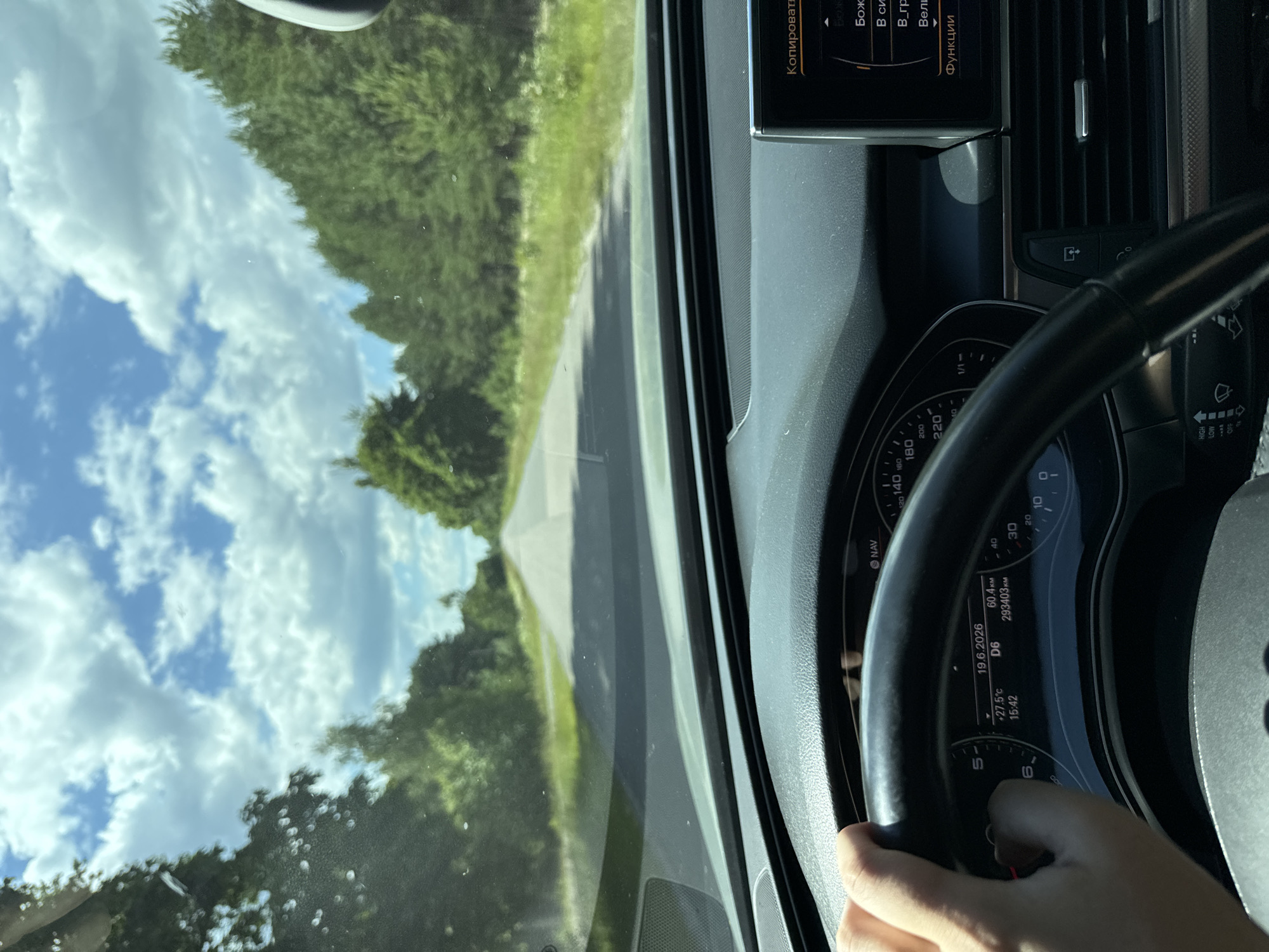 The image is a point-of-view shot from inside a car, looking forward along a paved road that cuts through a wooded area. The sky is blue with fluffy white clouds. The car's dashboard is visible in the foreground, with a hand on the steering wheel. The instrument cluster shows a date of "19.6.2026", a temperature of "+27.5°C", and an odometer reading of "293403 KM". A navigation screen is visible on the right side of the dashboard, displaying text in Cyrillic script. The scene suggests a journey on a sunny day through a rural or suburban environment, likely in Ukraine given the Cyrillic text. There is no indication of the city of Kostiantynivka, Ukraine in the image.