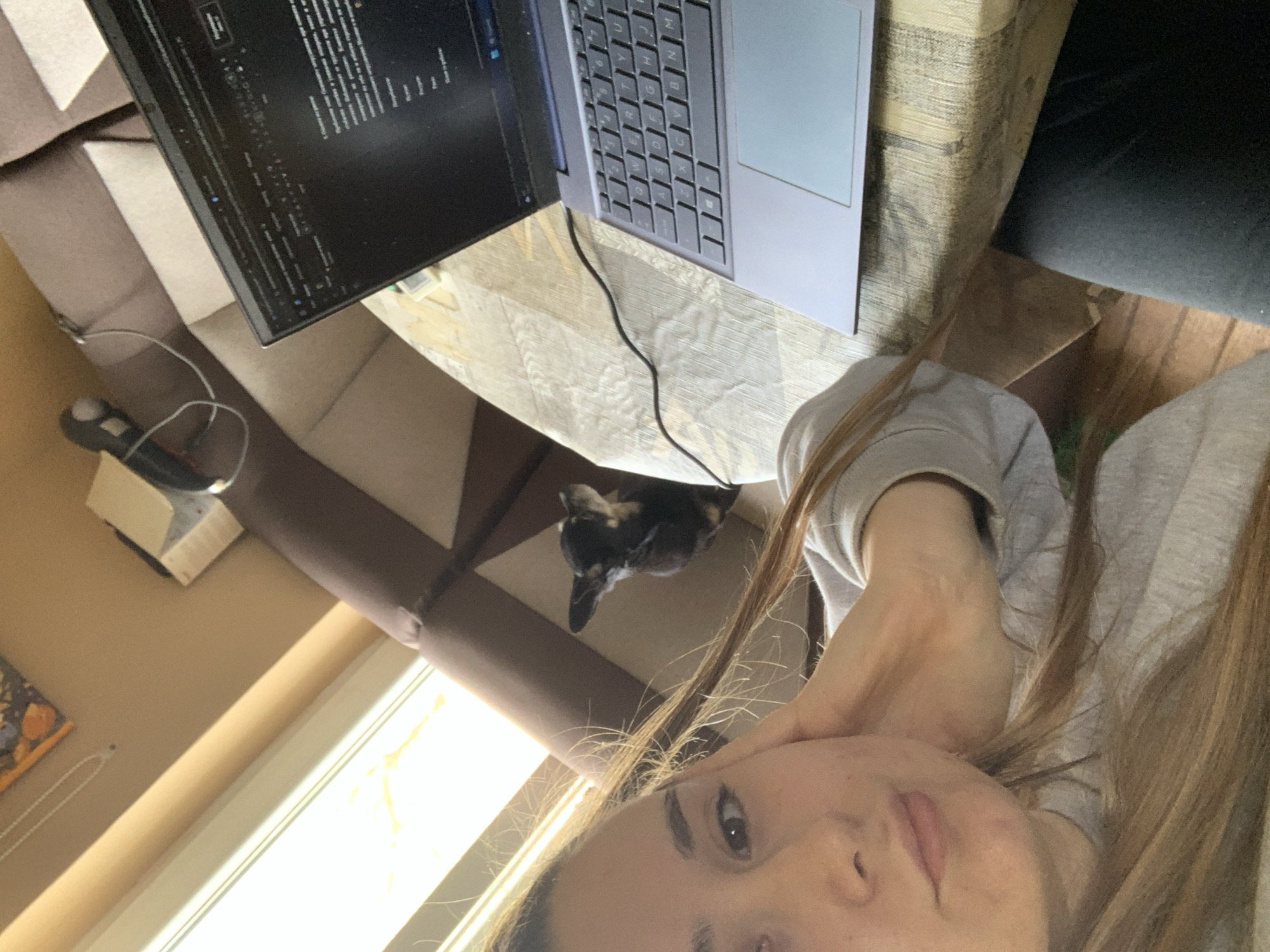 A young woman is taking a selfie while sitting at a desk with a laptop. A small dog stands on the floor by the desk. A monitor is connected to the laptop. A mouse and mousepad sit on a shelf to the left of the desk. A window with light shining through is visible in the lower left of the photo. No details are available to confirm the location in Lehchevo, Bulgaria.