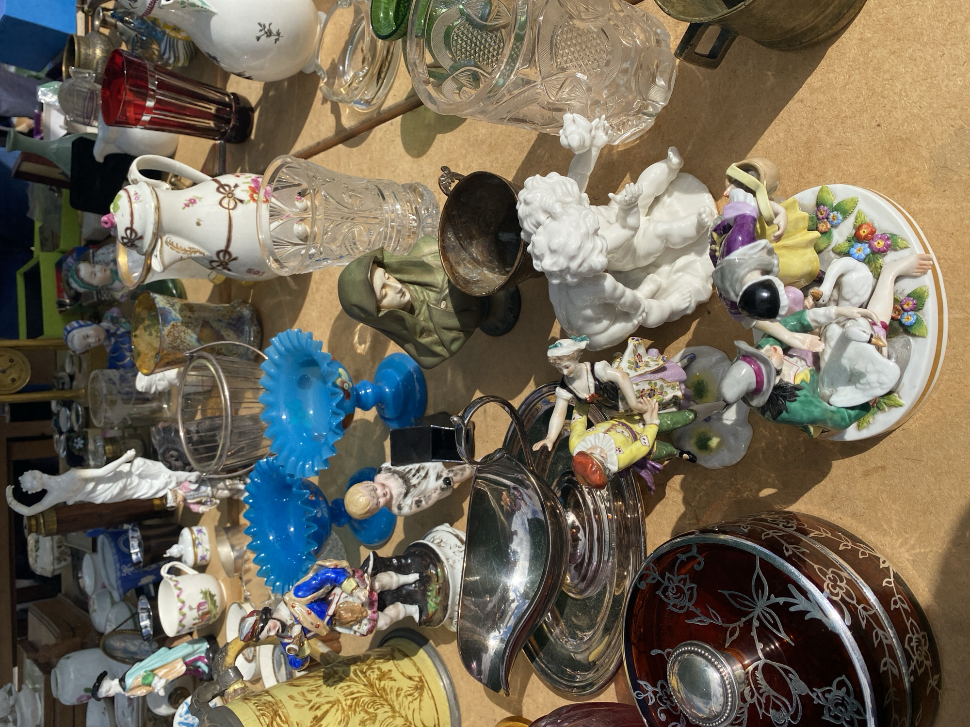 This is a close-up, overhead shot of various antique objects displayed on a wooden surface, likely at an outdoor market. The scene is bathed in bright sunlight, casting distinct shadows. In the foreground, to the left, sits a dark red, lidded glass jar with an ornate silver floral design. To its right, a group of porcelain figurines depicting people and a swan are arranged on a white base adorned with flowers. One figurine wears a yellow and purple outfit, another a green and yellow garment, and a third is topped with a grey hat. Further back, a silver gravy boat rests on a matching saucer, reflecting the light.
Behind these items, a collection of decorative glass pieces is visible, including two vibrant blue glass bowls with ruffled edges, a clear crystal glass with intricate cut patterns, and a tall, dark red glass vase. Several other ceramic and porcelain objects are scattered throughout the frame, including a small pitcher with floral decorations, figurines of people in historical attire, and busts. A white statue of a woman in a flowing robe is prominent in the background, near a tall wooden object that resembles a telescope. The overall impression is of a curated display of diverse antique collectibles. The specific location in the city of Vienna, Austria is not identifiable from the visual information alone.