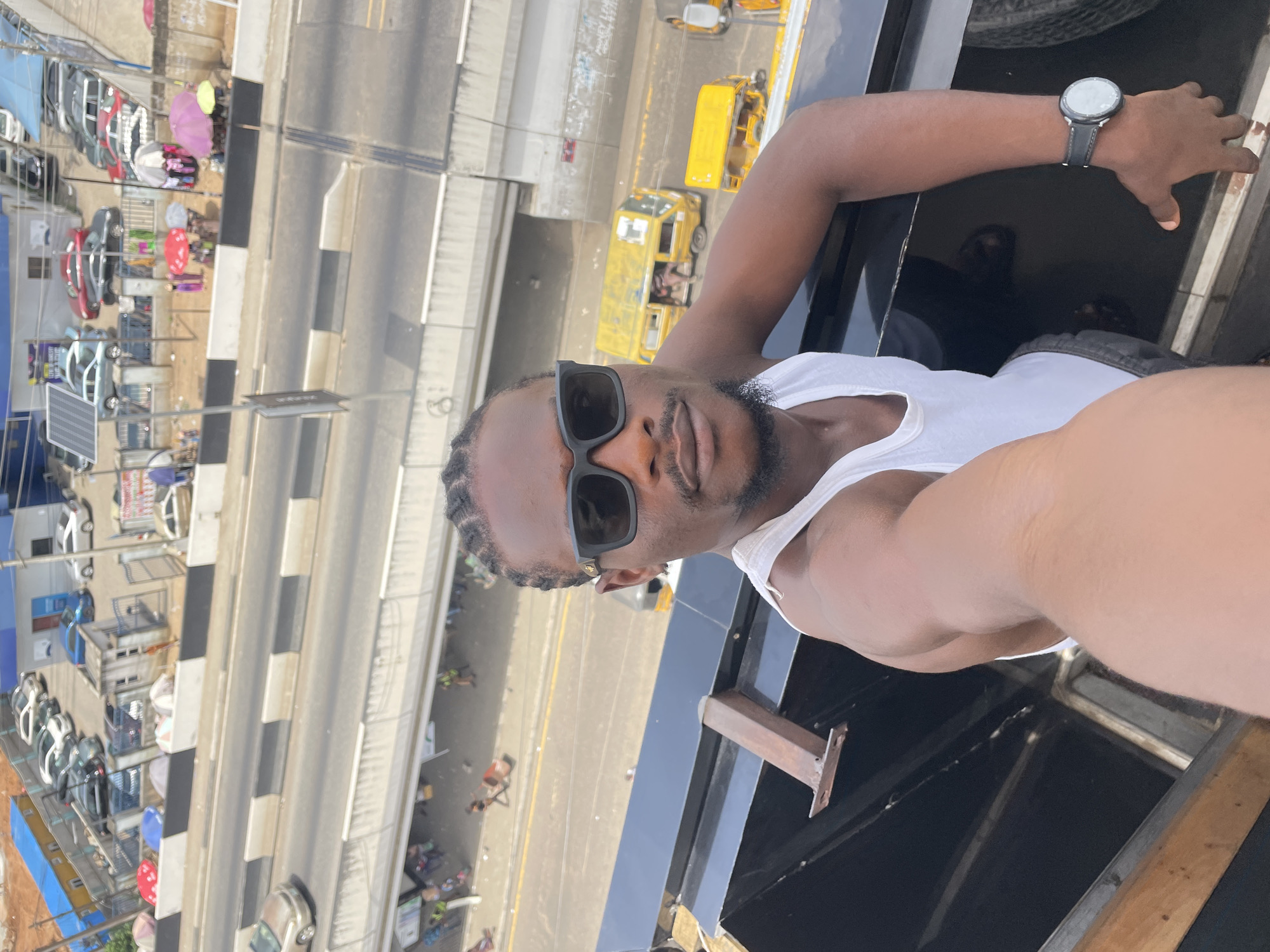A medium-shot, point-of-view photograph shows a Black man wearing sunglasses and a white tank top, standing on a high platform. He is wearing a wristwatch on his left wrist. Below the platform is a busy road. There are cars, buses, and other vehicles on the road, as well as people walking on the sidewalks. In the background, there are buildings and other structures. Based on visual cues such as yellow commercial buses (danfos) and architectural styles, the location is likely Ikeja, Nigeria.