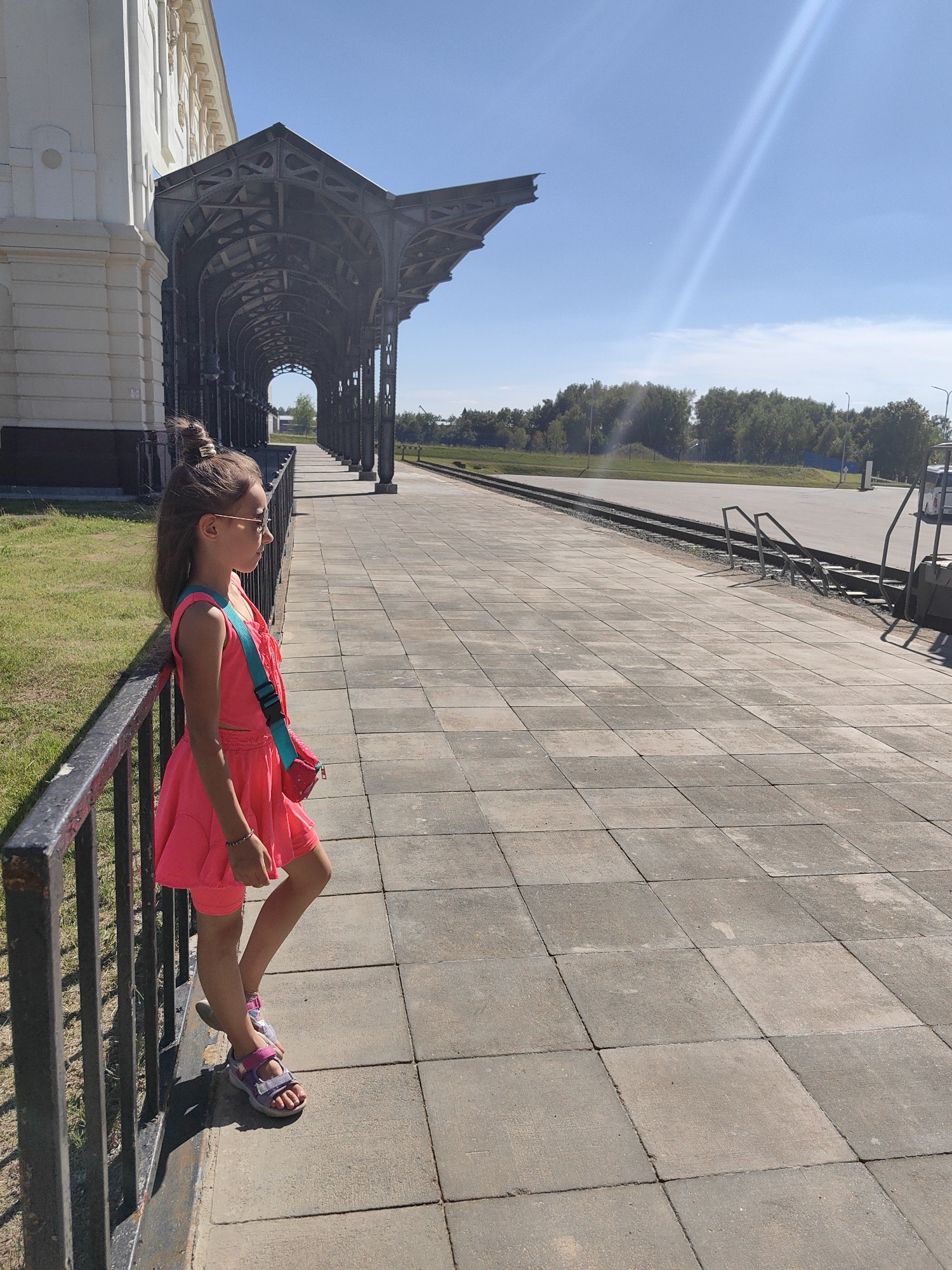 A young girl stands on a paved platform next to a black metal railing. She is wearing a bright pink dress and sandals, and has a turquoise crossbody bag. She is looking off to the right, with her hair tied up in a bun and wearing sunglasses. To her left is a large, ornate building with a white facade and a dark, arched metal awning extending over the platform. The platform continues into the distance, flanked by train tracks and bordered by trees and a clear blue sky. The sun is shining brightly, casting shadows on the ground. The scene appears to be an outdoor train station or a similar public space.