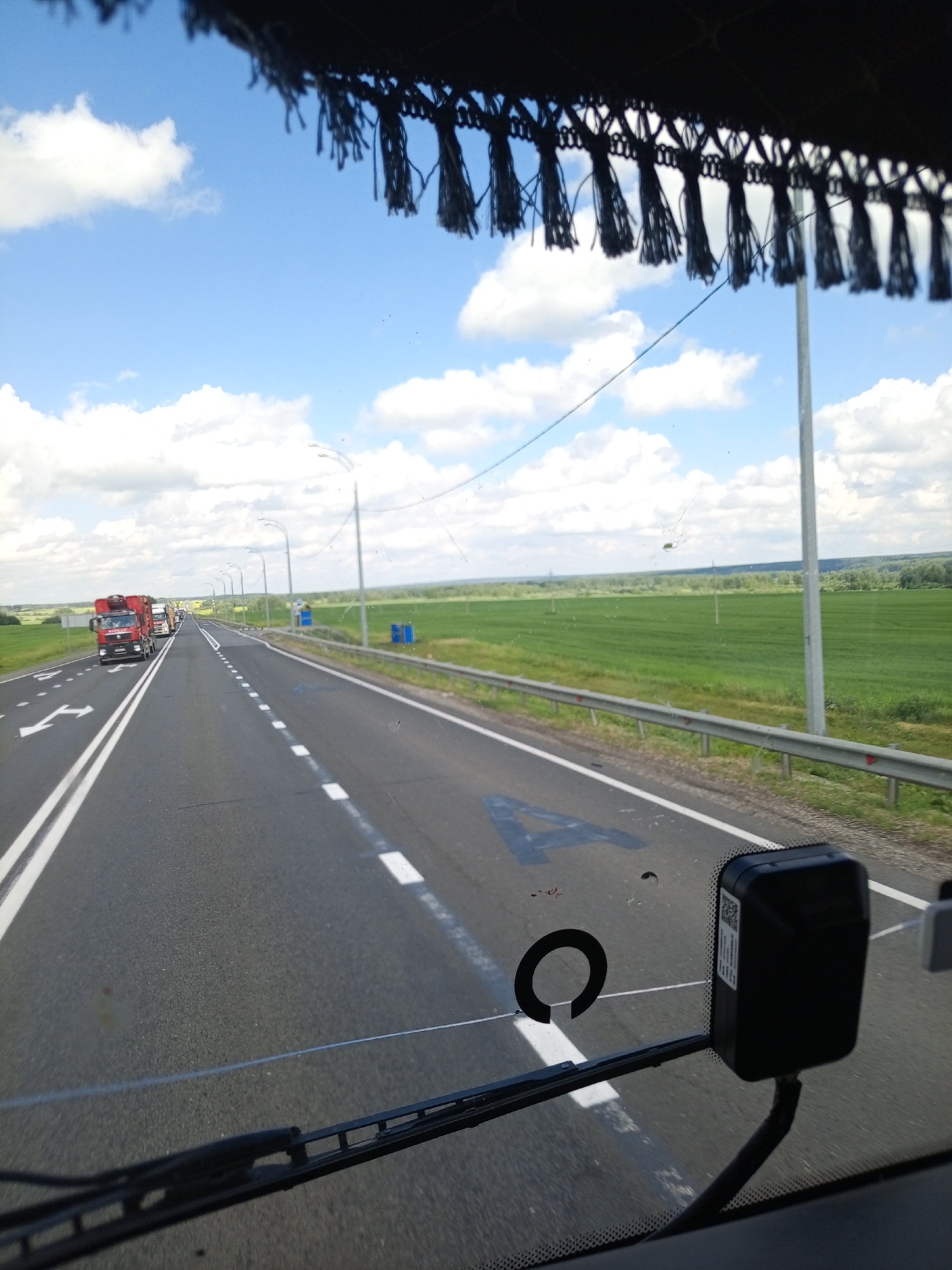 This media file captures a view from inside a vehicle, likely a truck cab, looking out through the windshield. The subject is a highway with several red trucks driving in a line. The road is asphalt with white lane markings, including directional arrows indicating a turn. The scene is set in a rural environment with lush green fields stretching out on either side of the road, bordered by a metal guardrail and a line of utility poles. In the distance, a line of trees marks the horizon. The sky is a bright blue with scattered white, fluffy clouds, suggesting it is daytime and the weather is fair. The light is bright, indicating it is likely midday. The image has a slight tint of green, possibly from the surrounding vegetation or a filter. There are some smudges and debris on the windshield, including what appears to be a large black letter "A" and a circular marking painted on the road itself, visible through the windshield. A black dashcam is mounted on the windshield. The overall impression is that of a long-distance journey on a clear summer day. There is no visible text in the image apart from the QR code and serial number on the dashcam.