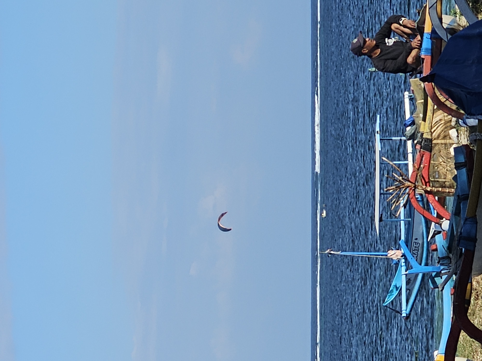 An eye-level medium shot captures a serene beach scene under a clear, blue sky. In the distance, a kiteboarder is actively engaged in their sport, their colorful kite soaring against the horizon. The ocean stretches out, a deep blue expanse broken by a line of white, breaking waves. Closer to shore, traditional outrigger boats are beached on the sand. One boat, painted a vibrant blue, has the word "MATE" visible on its side. A man, wearing a dark cap and long-sleeved shirt, is seated on another beached boat, looking out towards the ocean. He appears to be relaxing or observing the activity. The foreground is filled with the colorful hulls and wooden structures of several other outrigger boats, suggesting this is a popular spot for fishing or watersports. The overall atmosphere is peaceful and typical of a tropical coastal location.