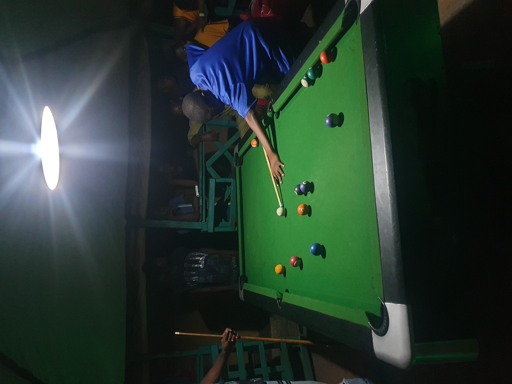 The image shows a group of people playing a game of billiards in what appears to be an indoor, dimly lit setting. The scene is dominated by a green billiards table, with several colored balls scattered across its surface and two cue sticks in use. One person, wearing a blue shirt, is in the process of striking a ball with a cue stick. The lighting is unusual, with a strong, bright light source on the left side of the image, casting a glare and long shadows. This suggests it might be evening or nighttime, and the light source could be a lamp or bulb. The background is dark and indistinct, but hints of wooden structures and other people are visible, suggesting a casual social gathering, possibly in a bar or recreational space. The overall atmosphere seems relaxed and focused on the game. There is no visible text in the image. The location is specified as Lokoja, Nigeria.