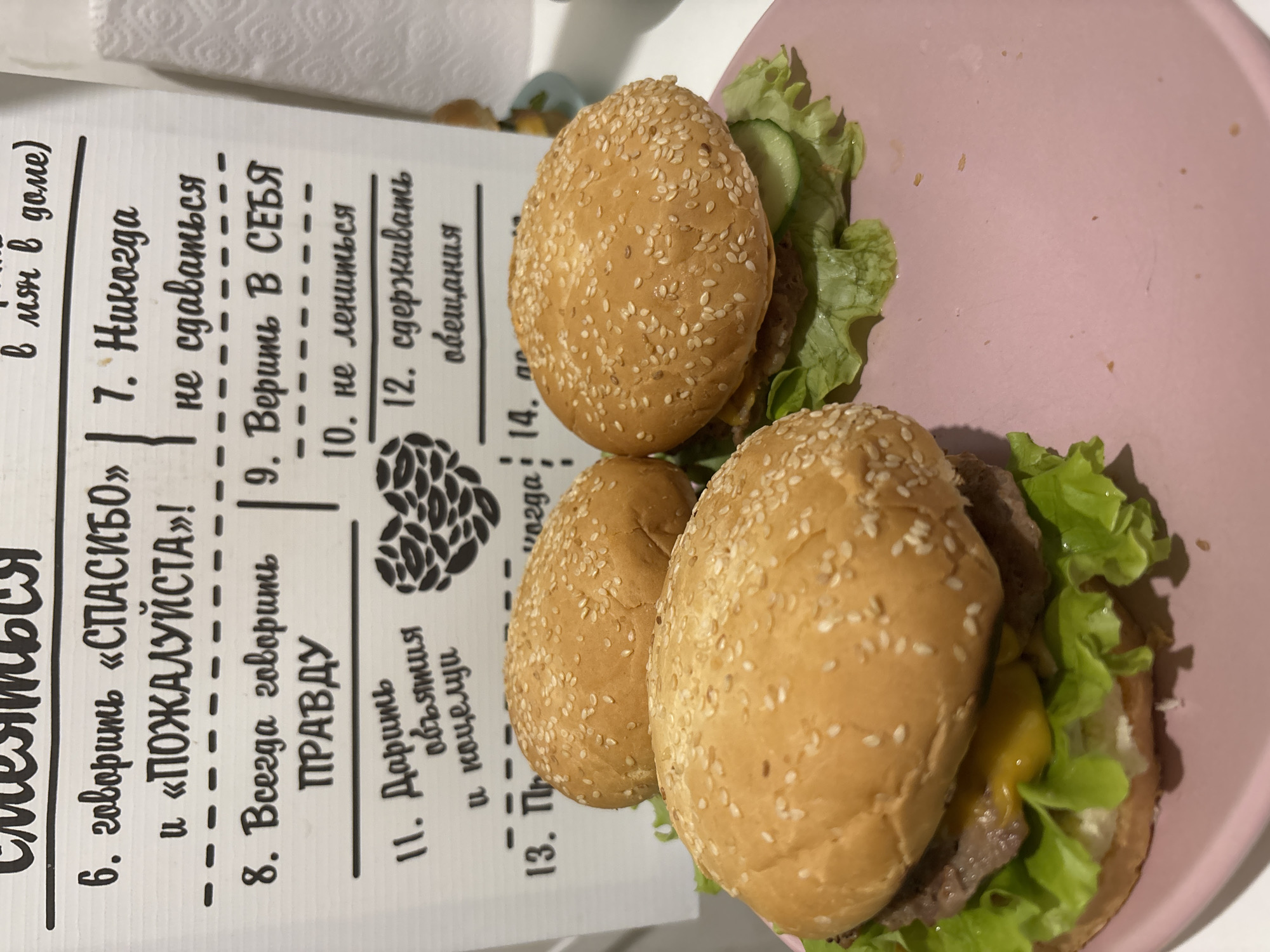 A close-up, slightly tilted shot shows three burgers on a pink plate next to a white paper with black text written in Russian. The burgers have sesame seed buns and appear to contain lettuce and a beef patty. The paper lists rules or affirmations, including phrases like "Say 'Thank you' and 'Please,'" "Never give up," and "Believe in yourself." The image was taken in Chaikovsky, Russia.