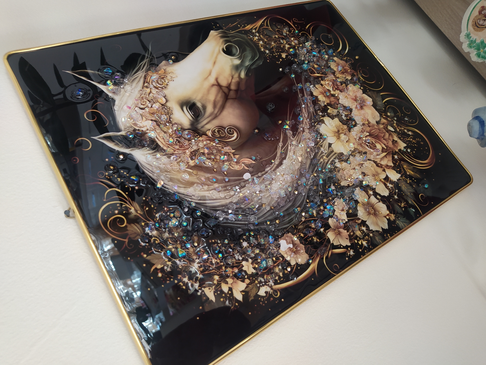 The image shows a framed artwork of a horse's head with a crown of flowers and jewels, rendered in a style that incorporates both painting and 3D elements. The horse is depicted with a light-colored mane and face, adorned with intricate floral and sparkling embellishments. The background is dark, with gold swirling accents and clusters of dark and iridescent crystals or gems. The artwork has a glossy finish and is framed with a thin gold border. The overall impression is one of opulence and fantasy. The image appears to be taken indoors, with a white wall visible in the background, suggesting it is displayed in a home or gallery setting. There is no discernible indication of the city of Livorno, Italy, within the image itself. The image is a close-up, angled shot of the artwork.