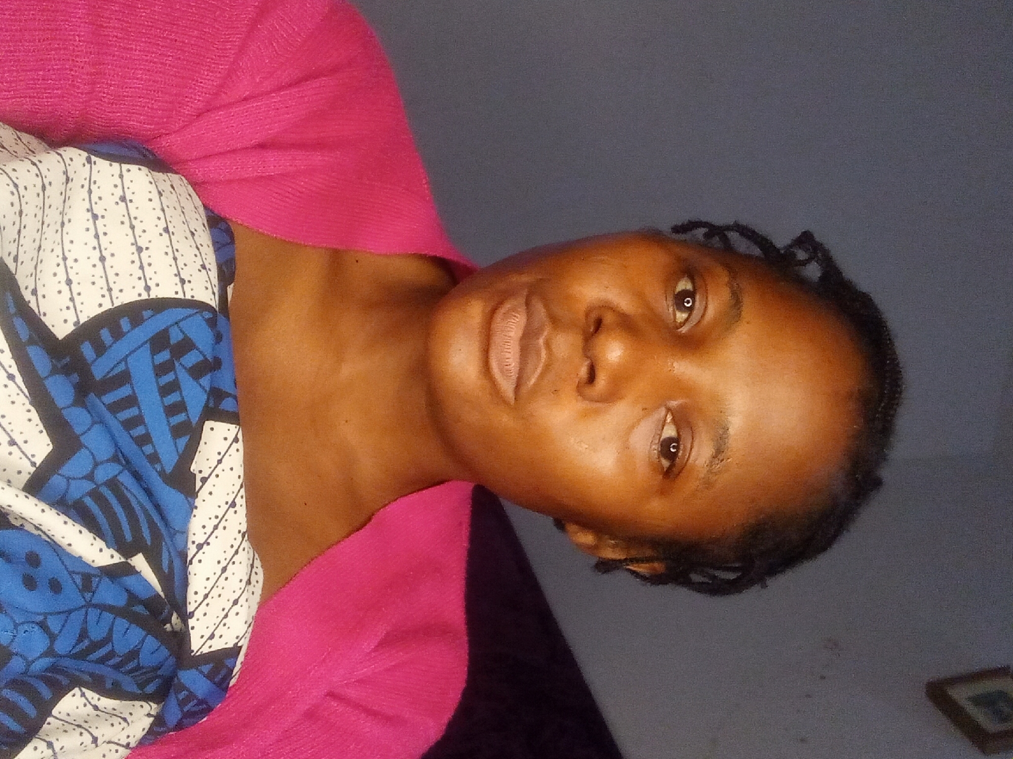 A medium shot photo shows a Black woman from the chest up. She has dark braided hair and is looking directly at the camera. Her lips are parted, and she is wearing a light pink sweater over a blue and white patterned top. The background is a dark gray wall with a framed picture on the upper right side.