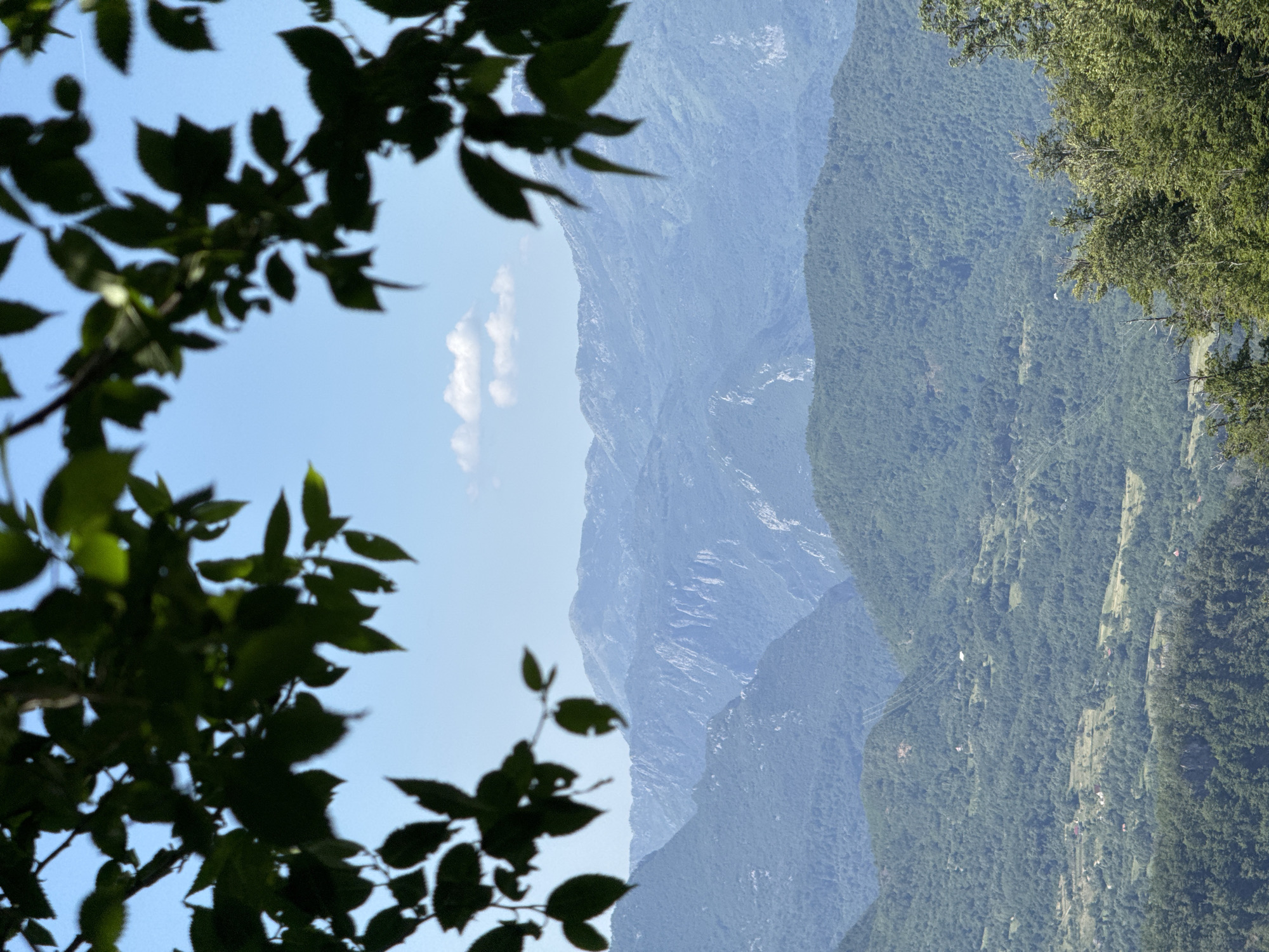 An outdoor, eye-level, vertical shot captures a view of distant, hazy blue mountains under a clear, pale blue sky. The foreground is framed by dark green, out-of-focus leaves from a tree branch. The mountains appear layered, with the closest ones being lush green and speckled with small buildings, suggesting a rural or village area. Further into the background, the mountains become progressively bluer and less defined, with jagged peaks and lighter patches indicating rock faces. A few scattered, wispy white clouds are visible in the sky. The scene conveys a sense of natural beauty and vastness.