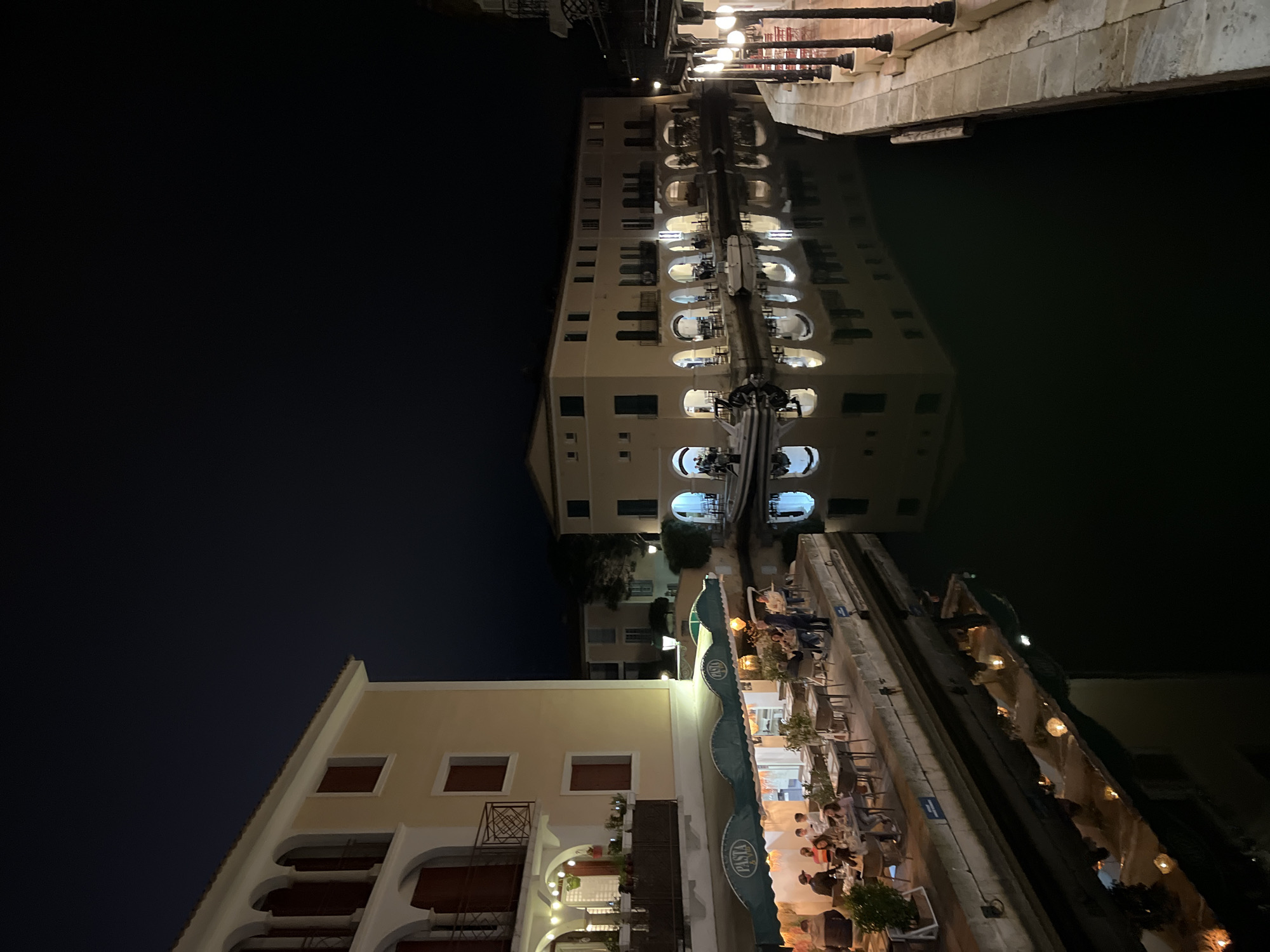 The image depicts a nighttime scene along a canal in Grimaud, France. Buildings are visible on both sides of the canal, with lights reflecting in the dark water. People are seated at outdoor tables alongside the canal, possibly dining at restaurants. The architecture appears to be a mix of residential and commercial, with archways and balconies visible. The overall atmosphere suggests a relaxed, tourist-oriented setting.