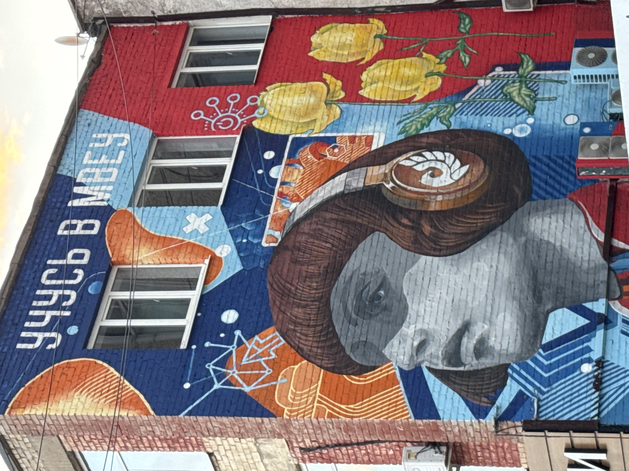 This is a photograph of a mural painted on the side of a multi-story brick building in Izhevsk, Russia. The mural depicts a stylized portrait of a person wearing headphones, rendered in shades of gray, with brown hair. To the right of the portrait are three yellow flowers with green stems and leaves. The background of the mural is a vibrant mix of red, blue, and orange, with geometric patterns and abstract shapes. White text in a Cyrillic font appears at the top of the mural, reading "УЧУСЬ В МВЕУ" which translates to "I am studying at MVEU." The building has several windows, some of which are visible through the mural. Air conditioning units are mounted on the exterior walls, and electrical wires are visible overhead. The mural covers a significant portion of the building's facade, suggesting it is a public artwork.