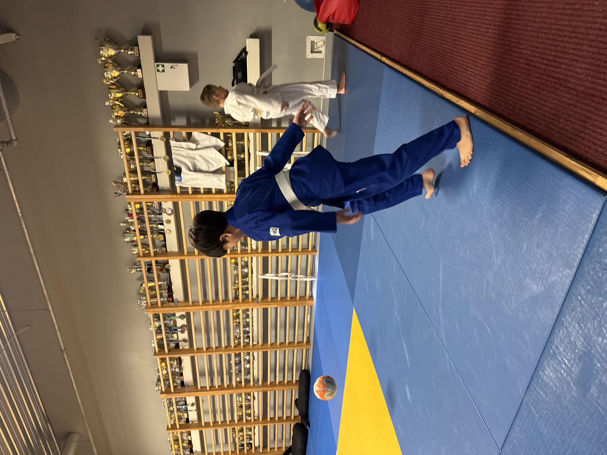 The image depicts an indoor martial arts training facility, likely a dojo, in Tallinn, Estonia. In the foreground, a young person wearing a blue judo gi is visible from the waist down, with their feet on a blue mat. A multi-colored ball is in mid-air to the left of the person. In the mid-ground, another young person in a white judo gi is walking away from the camera. The background features a wall lined with wooden shelves displaying numerous trophies and medals. Two dark, elongated bags are placed on the floor near the shelves on the left side of the image. A first-aid kit is mounted on the wall on the right side, above a shelf holding some bags. The floor is covered with a blue mat, transitioning to a yellow mat in the upper left corner, and a red carpet along the right edge of the frame. The lighting suggests an indoor setting with overhead fluorescent lights.