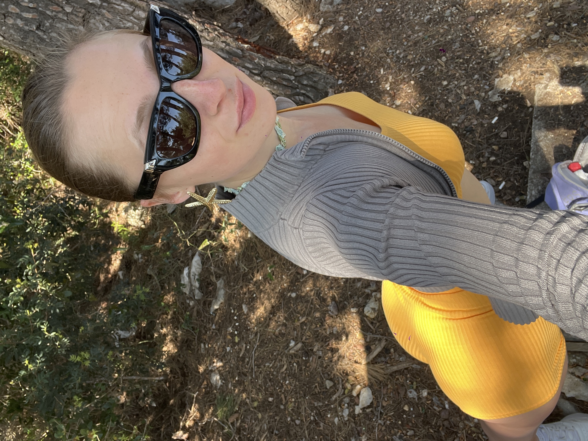 A woman is seen from an overhead angle, standing outdoors in what appears to be a natural environment. She is wearing sunglasses, a gray knit zip-up sweater over a yellow top and skirt, and earrings in the shape of starfish. The ground is covered with dry leaves and twigs, indicative of a wooded or park-like setting. The scene is illuminated by sunlight, casting shadows on the ground. Based on the vegetation and style, this image could be taken in a park or semi-natural area in Budva, Montenegro. However, without additional landmarks or contextual information, a precise location within Budva cannot be determined.