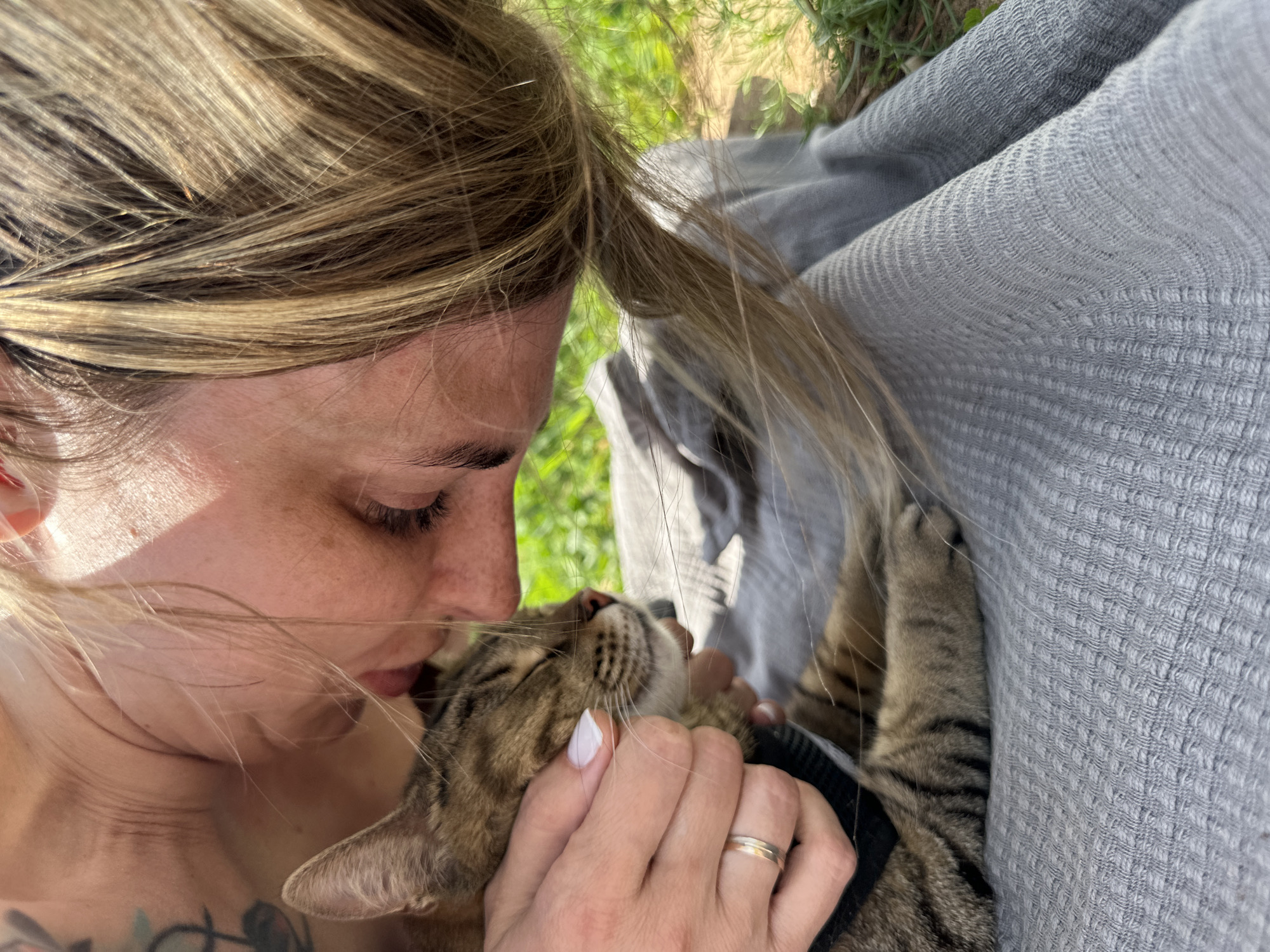 A woman with blonde hair is holding and kissing a tabby cat. The cat has its eyes closed. The woman is wearing a gold ring. The background includes a gray blanket or fabric and green foliage. The location and city are not discernible from the image.