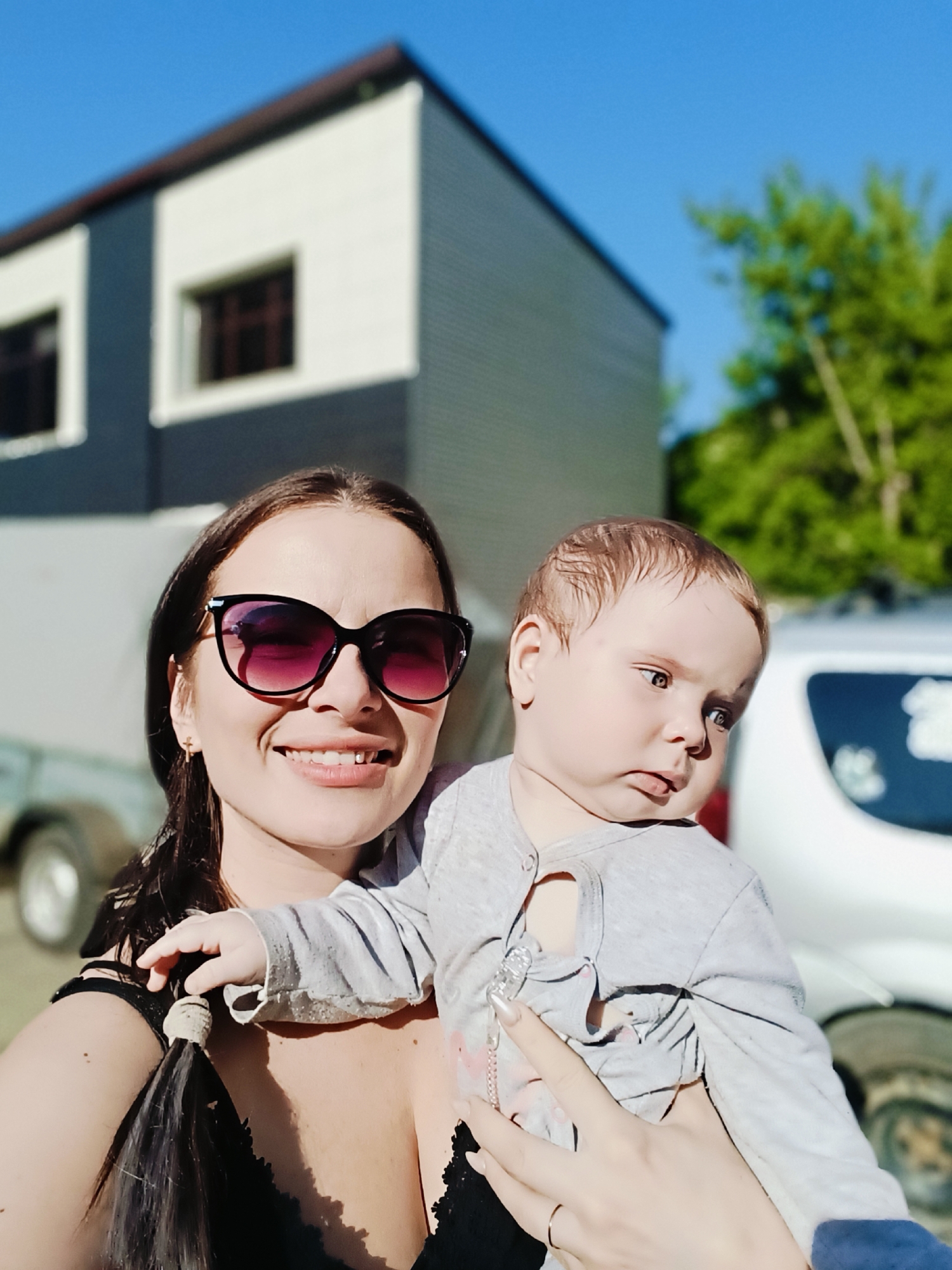 A woman wearing sunglasses holds a baby outdoors in Chaikovsky, Russia. The woman is smiling, has long brown hair pulled back in a ponytail, and is wearing a black top. The baby is wearing a gray onesie and looks to the side. Behind them is a light gray building with white accents and a brown roof, along with a silver trailer. Green trees are visible in the background, under a clear blue sky.