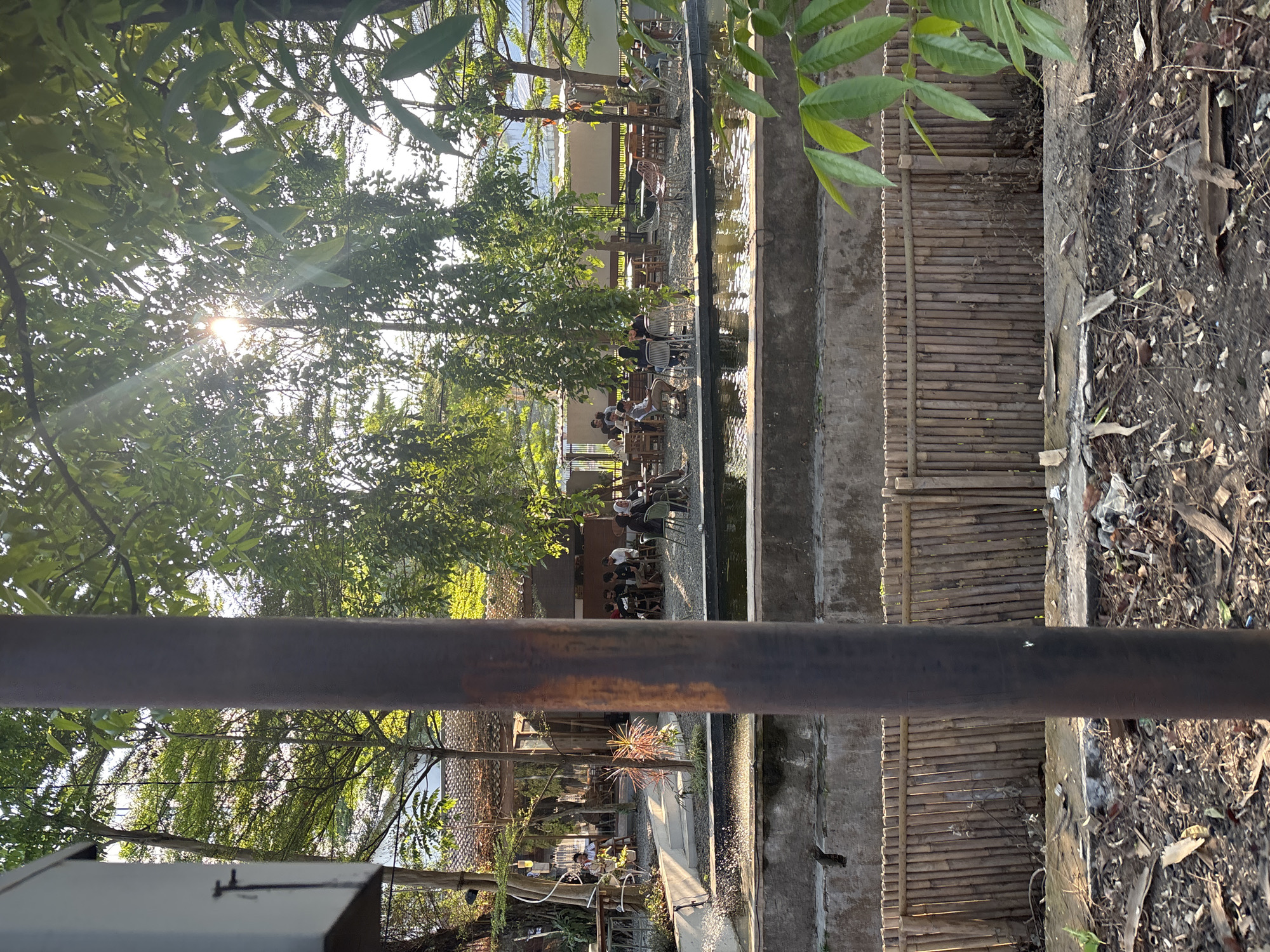 The image captures an outdoor seating area of a cafe or restaurant, possibly located in Cirebon, Indonesia, given the context. The scene is bathed in warm, late afternoon sunlight filtering through lush green trees. In the foreground, a thick, dark brown pole partially obscures the view. Behind it, a body of water, perhaps a pond or a small canal, reflects the sunlight and the surrounding greenery. A rustic fence made of vertical bamboo poles separates the water feature from a patch of dry, leaf-strewn ground with some scattered debris.
Further back, the seating area is set up with several tables and chairs, occupied by a number of patrons. Some individuals are seated in groups, engaged in conversation, while others appear to be enjoying their meals or drinks. The architecture of the establishment features a pitched roof with terracotta tiles and open-air seating areas, suggesting a casual, tropical ambiance. The presence of multiple dining tables and chairs indicates a commercial establishment designed for public gathering. The overall atmosphere is relaxed, with people enjoying the outdoor environment.