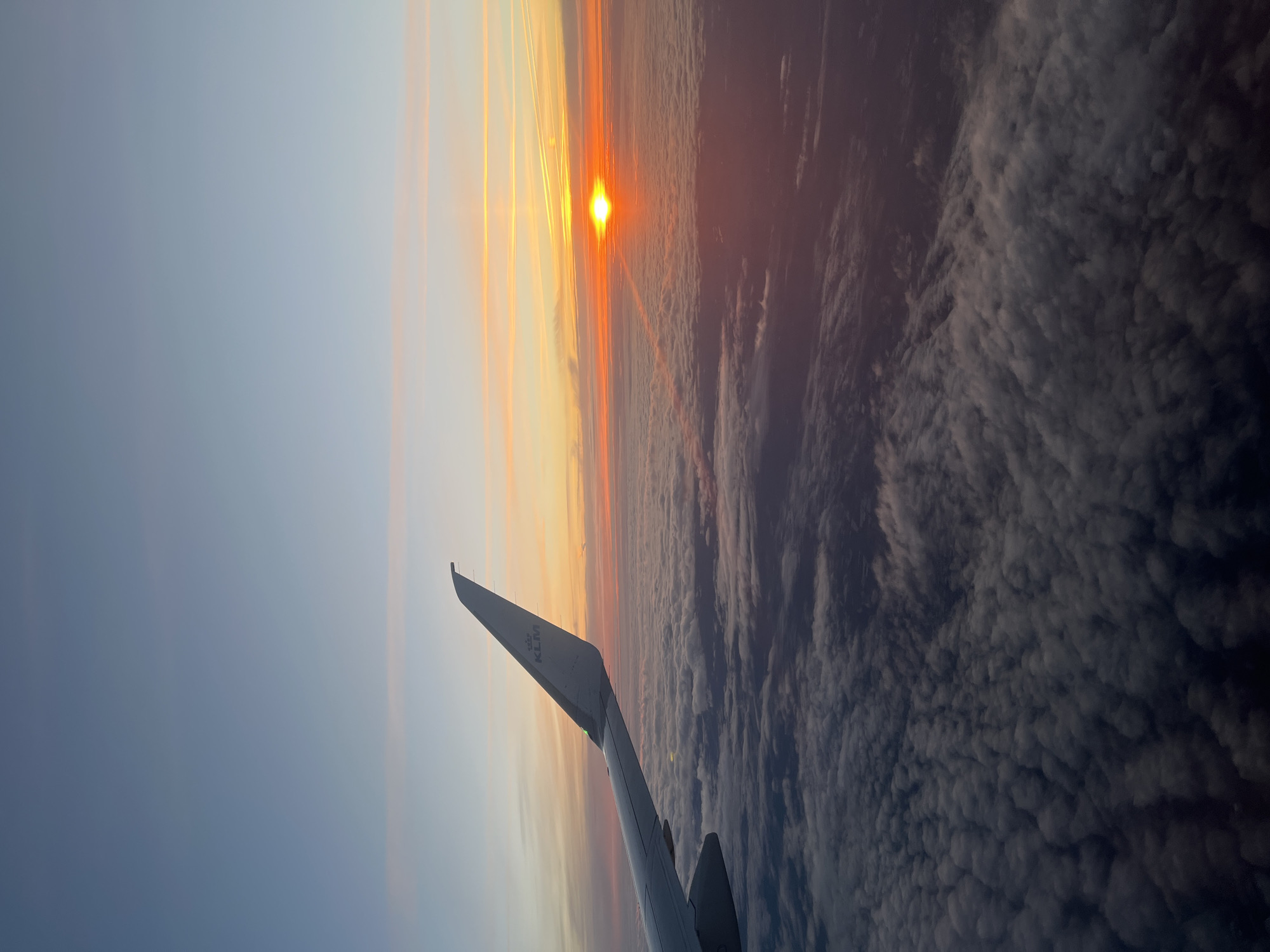 The image is a view from an airplane at sunset, possibly taken near Wouwse Plantage in the Netherlands given the presence of KLM (Royal Dutch Airlines). The wing of the airplane is visible in the lower left corner, bearing the KLM logo. The sun is positioned on the horizon, displaying a reddish-orange glow, and is partially obscured by layers of clouds. The sky transitions from a lighter blue near the top left to orange and red near the horizon. Below the horizon, a dense layer of gray and white clouds stretches out, resembling a landscape. The image is rotated slightly.