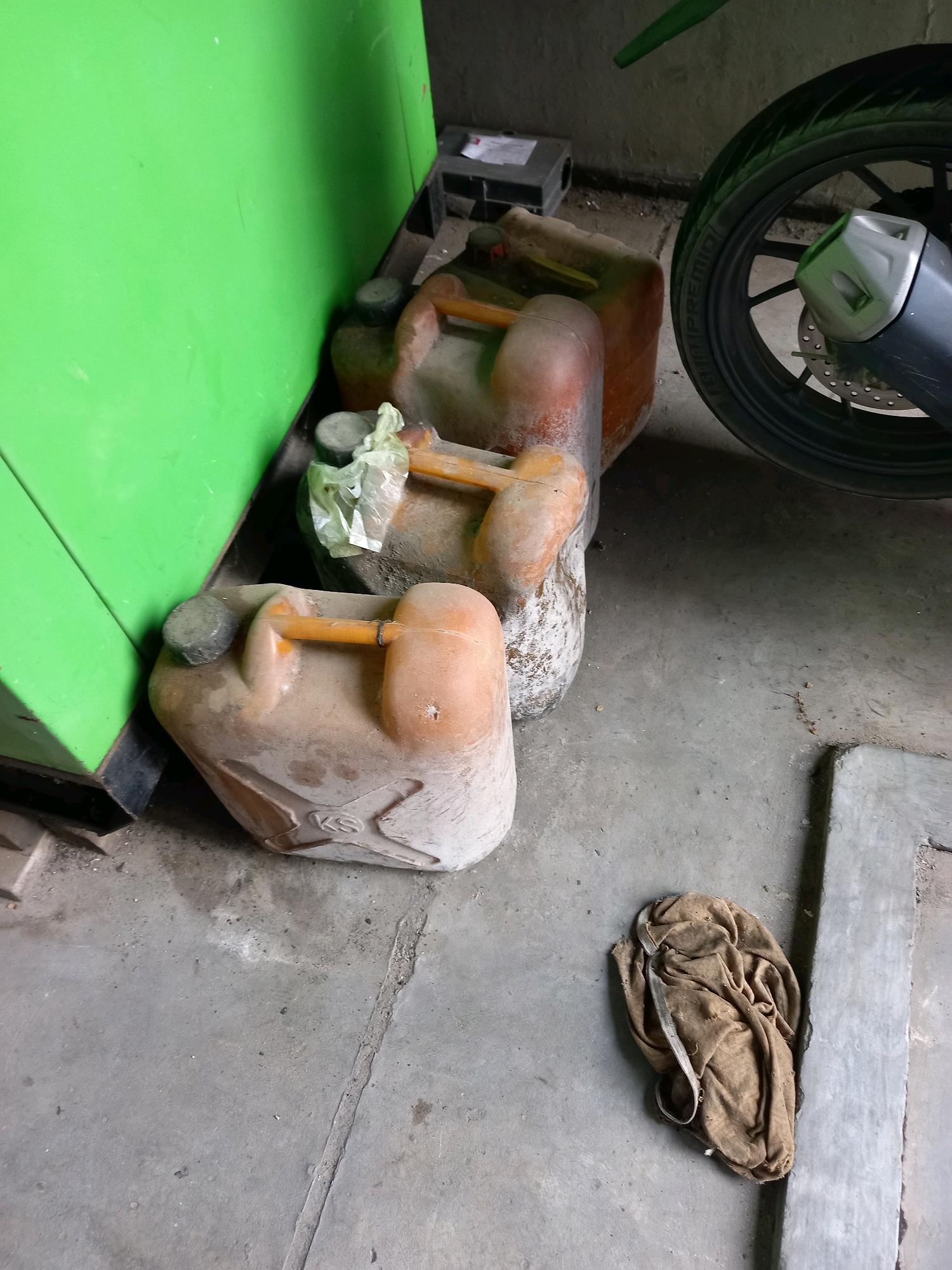 In an outdoor area in Samarinda, Indonesia, three orange plastic jerrycans are stacked on a concrete floor. The jerrycans appear to be empty and are dusty. A brown cloth is crumpled on the floor to the right of the jerrycans. A motorcycle wheel is visible in the upper right portion of the image. A bright green wall is to the left of the jerrycans.