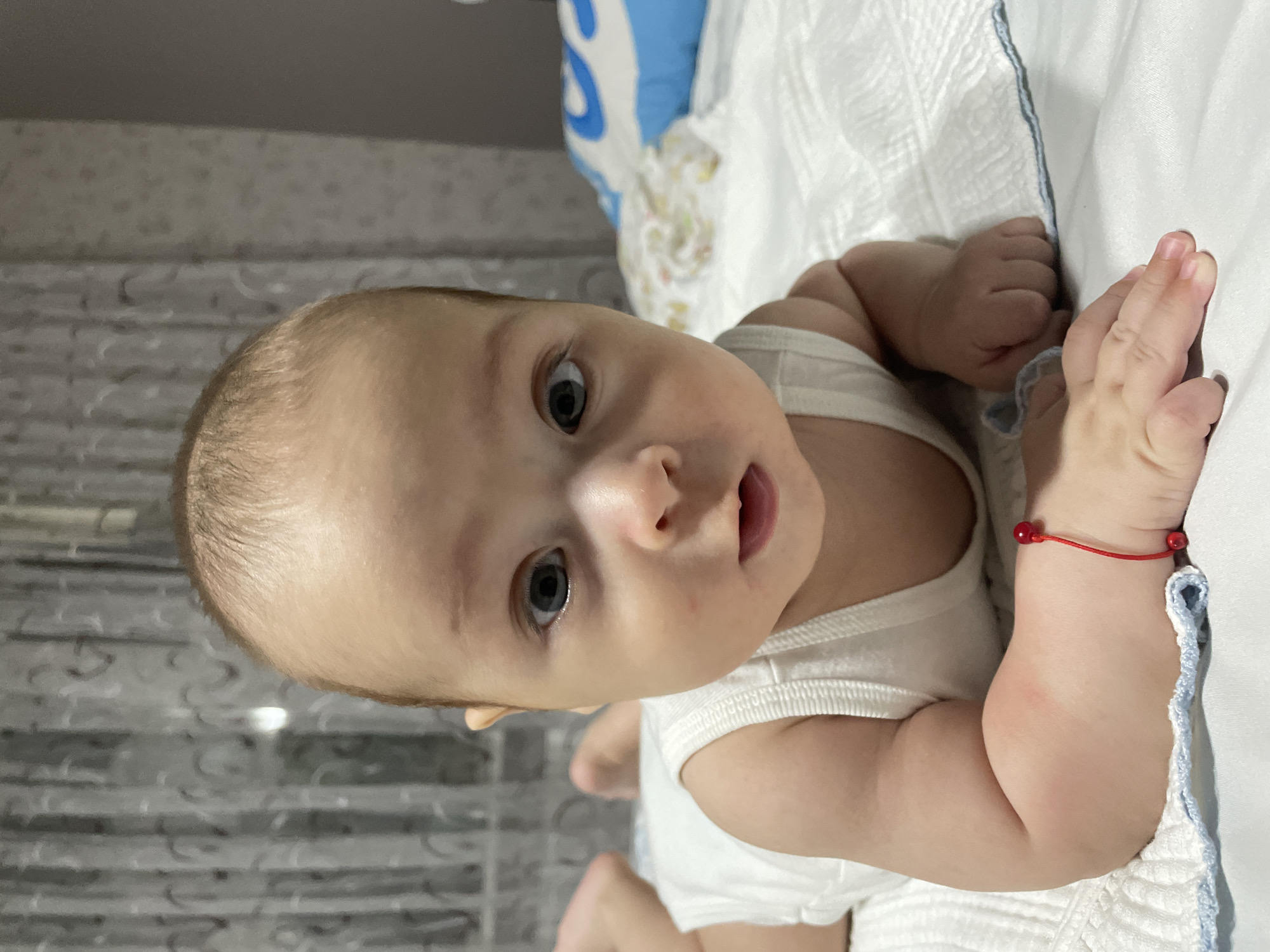 A close-up, eye-level shot shows a baby lying on its stomach on a white, textured surface, likely a bed. The baby is wearing a white undershirt. Its face is turned towards the camera, with large, dark eyes and a slightly open mouth. A thin red string bracelet with two small red beads is visible on its left wrist. In the background, sheer curtains with a patterned design are visible, and a blue and white pillow lies to the right of the baby. The lighting is soft, suggesting an indoor setting. There is no indication of the specific location in Fălești, Moldova.
