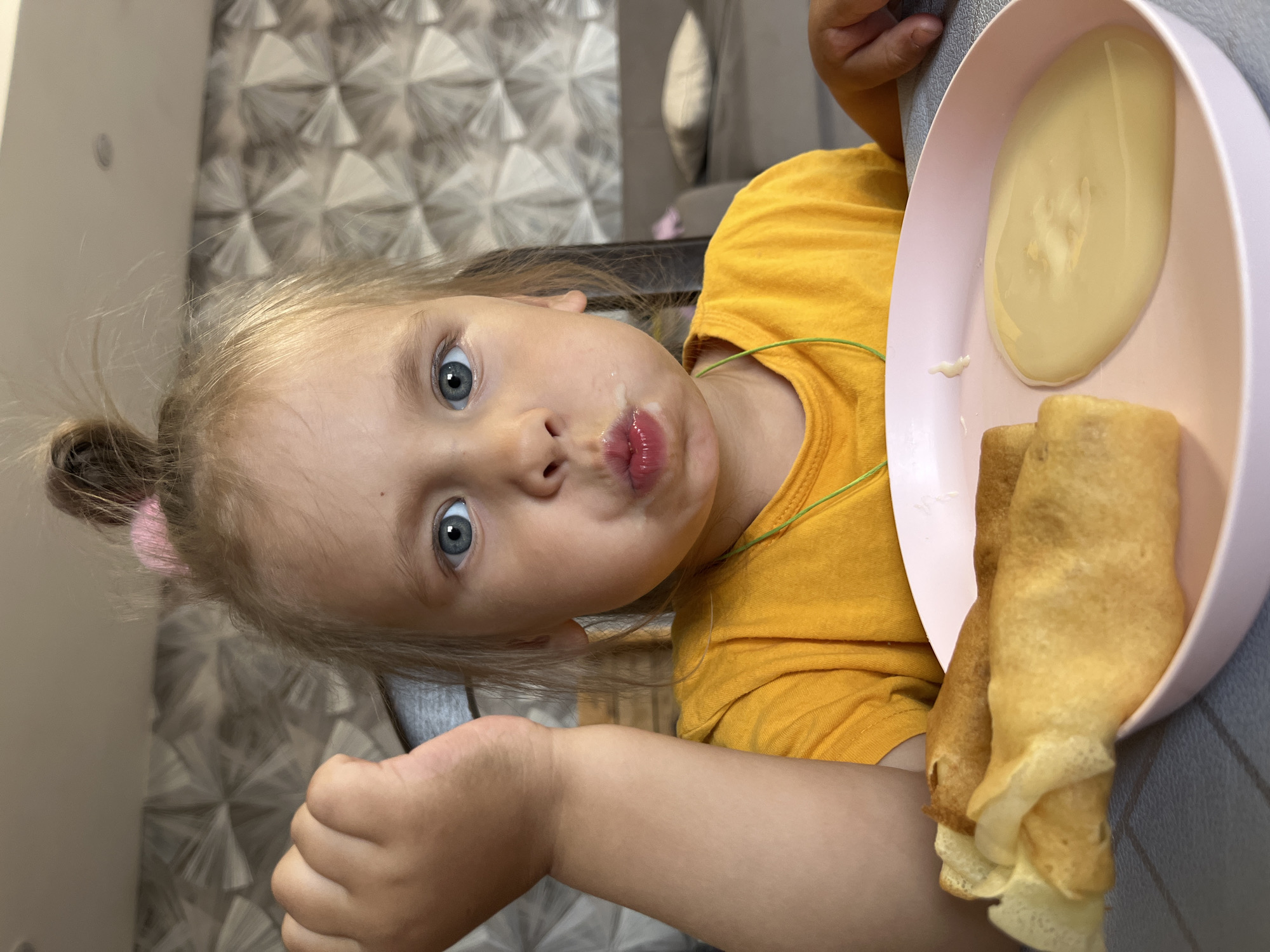A young girl with blonde hair pulled into a messy bun is sitting at a table, looking at the camera with wide, blue eyes. She has food around her mouth and is puckering her lips. She is wearing a yellow t-shirt. In front of her is a pink plate with a portion of yellow liquid, possibly syrup or a sauce, and two rolled-up crepes. The background shows a wall with a patterned wallpaper and a hint of furniture, suggesting an indoor setting, likely a home. The lighting is bright, indicating it could be daytime. The girl's expression is curious and perhaps a little mischievous. No text is visible in the image. The overall scene is casual and domestic, likely a mealtime for the child.