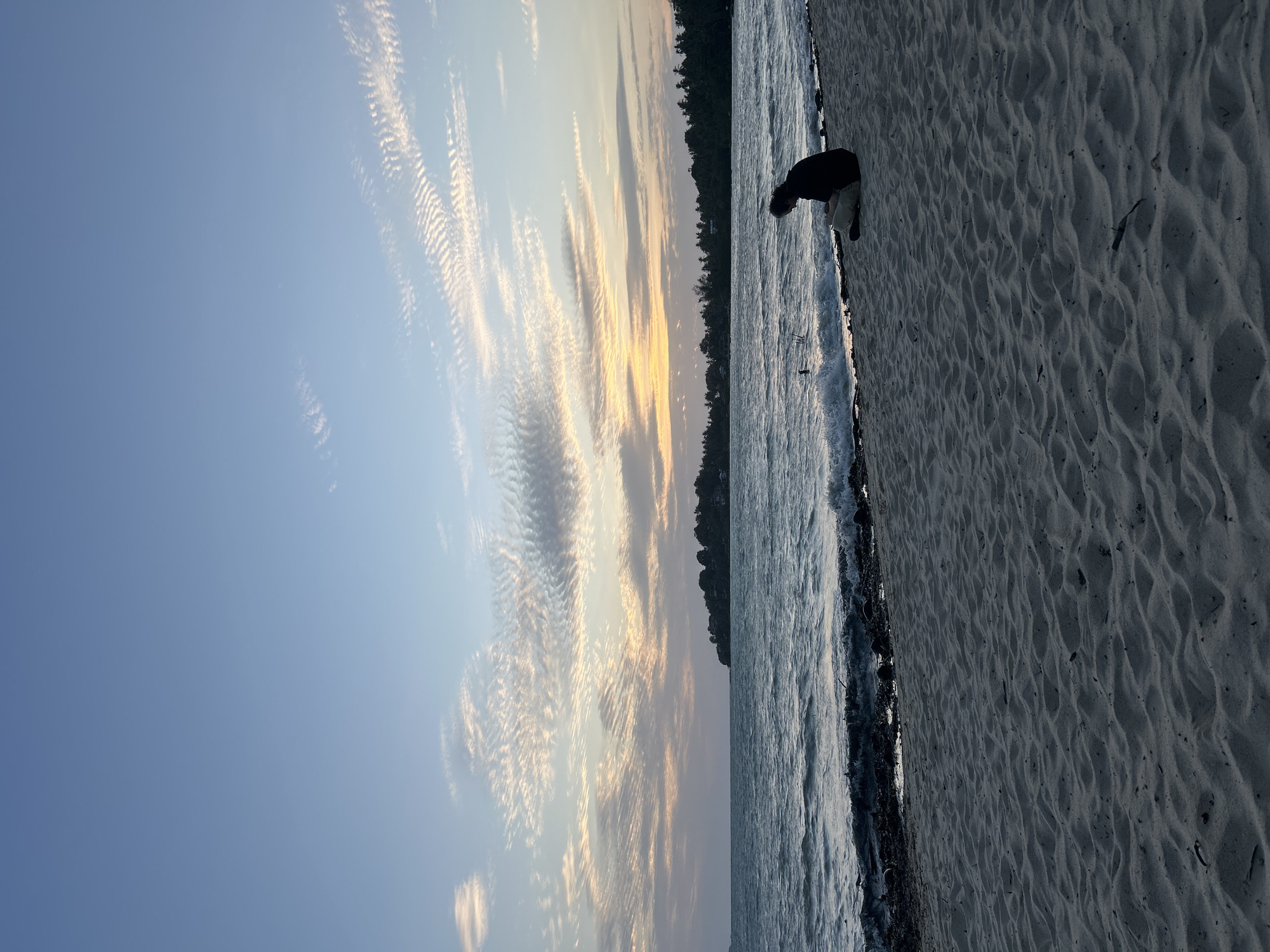 A person sits on a sandy beach in Ko Pha-ngan District, Thailand, facing the ocean at sunset. The sky is a soft blue with wispy, striated clouds illuminated by the setting sun, which casts a warm glow on the horizon. The ocean is a deep blue with gentle waves lapping at the shore. A line of dark green trees and some houses are visible along the distant coastline. The foreground is dominated by sand dunes and scattered debris.